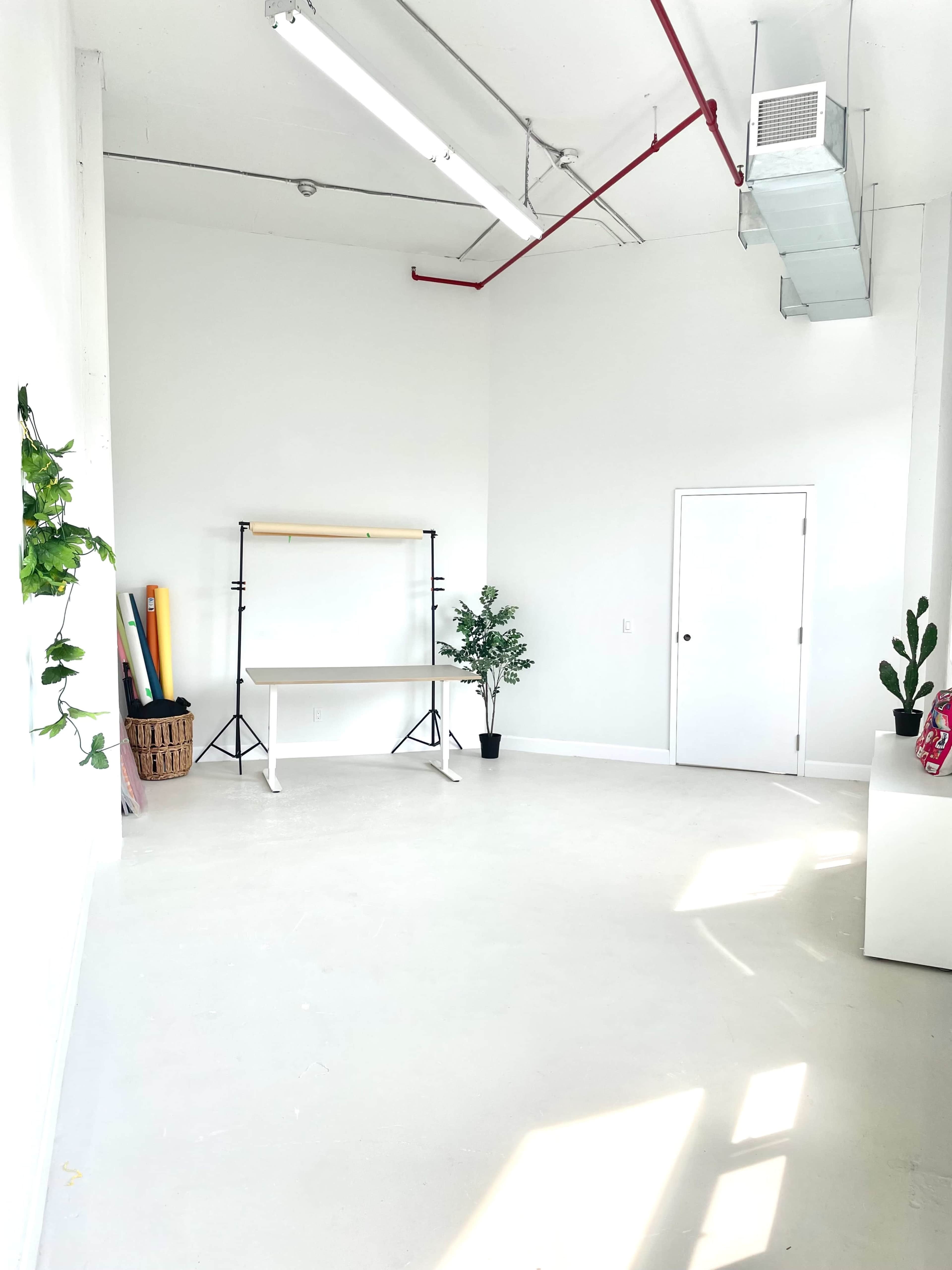 The image shows a spacious, bright studio with white walls, a table on a light-colored floor, and minimalist decor including plants and rolls of colored materials.