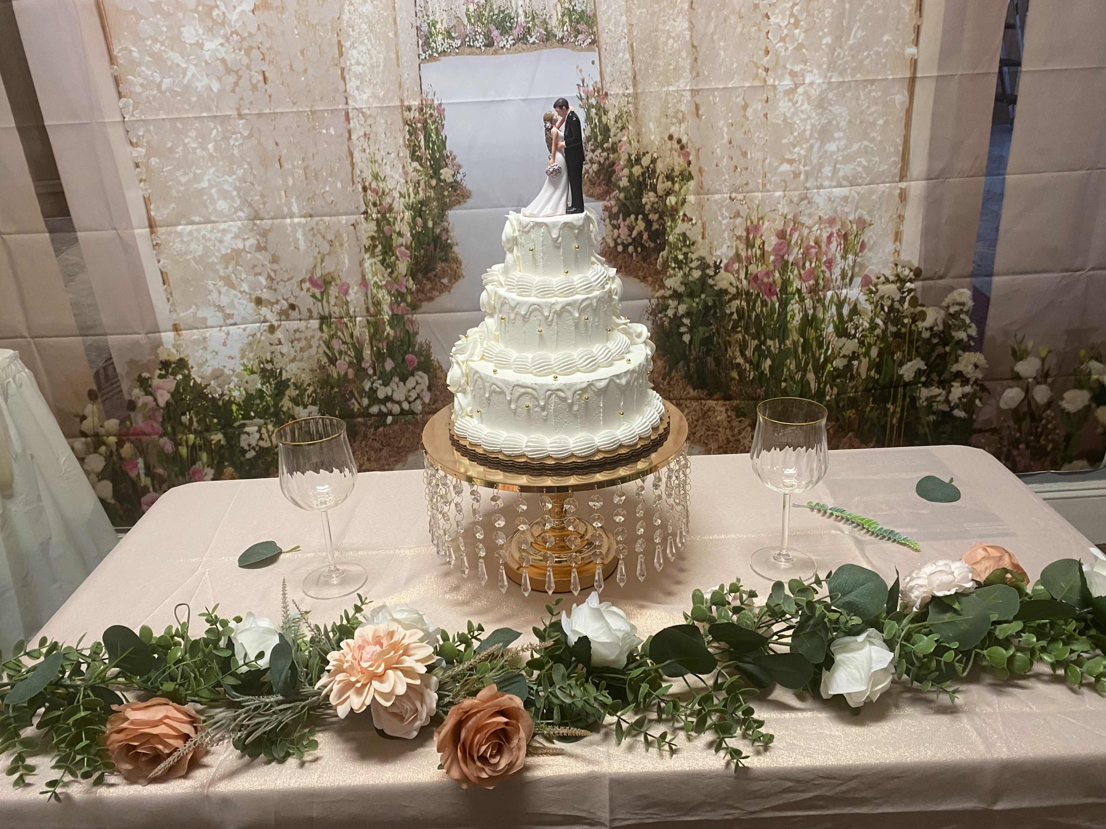 A three-tier wedding cake is displayed on a table adorned with greenery and flowers, set against a backdrop of a floral archway.