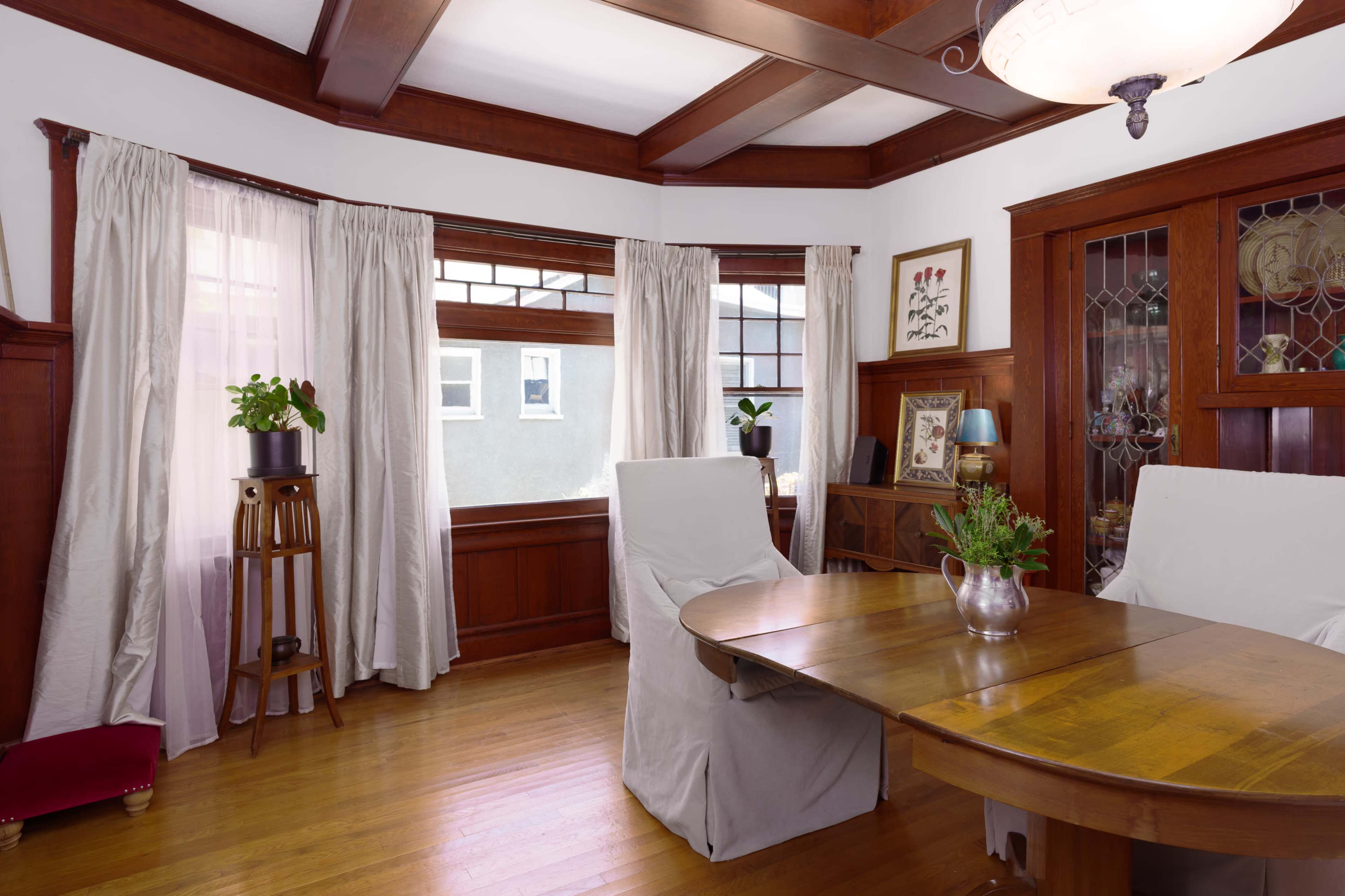 The image shows a warmly lit dining room featuring a round wooden table, four chairs covered with white slipcovers, and large windows with light-colored curtains.