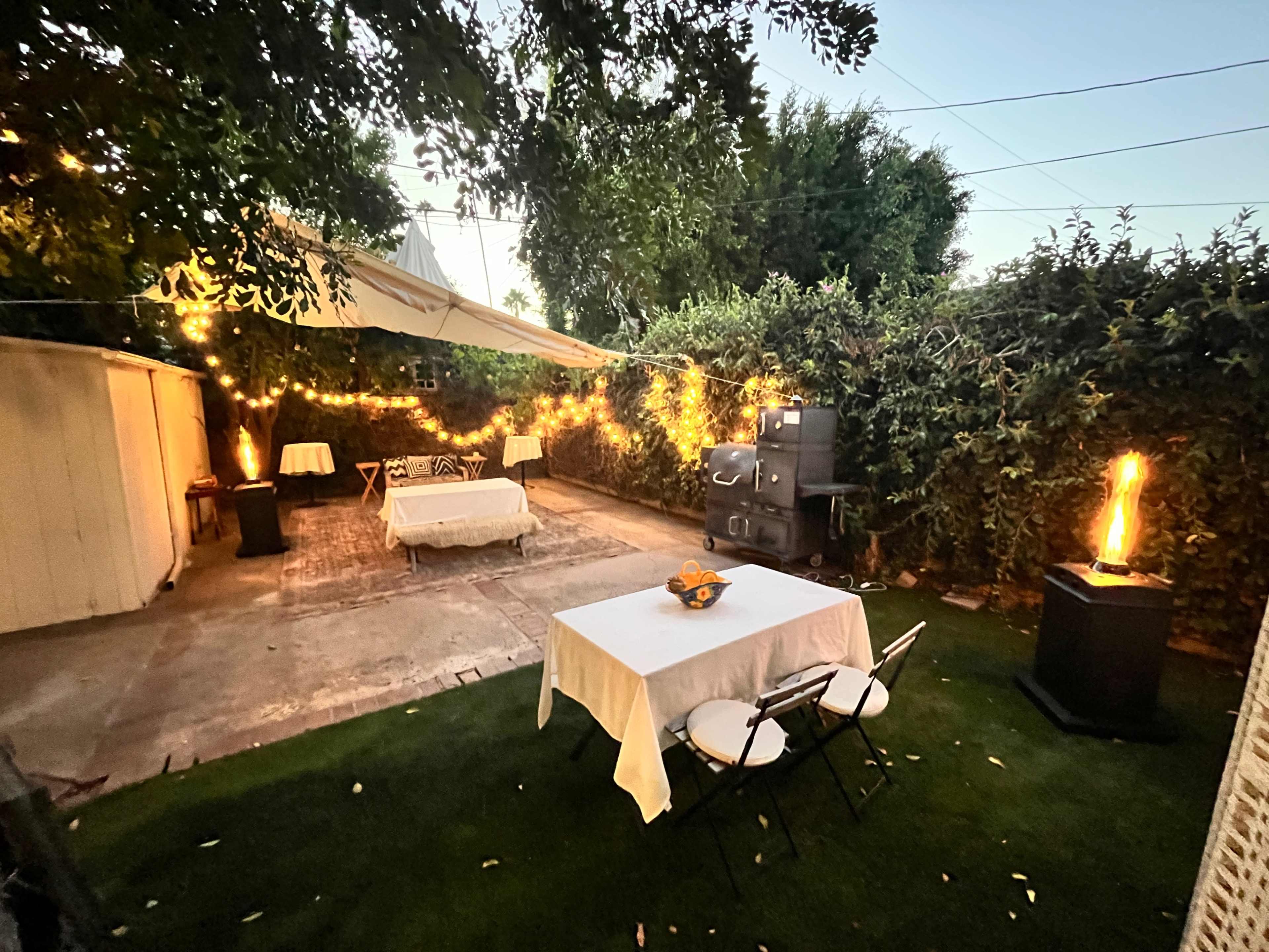 The scene shows a backyard patio with two tables covered in white cloths, surrounded by lush greenery and soft lighting from string lights in the evening.
