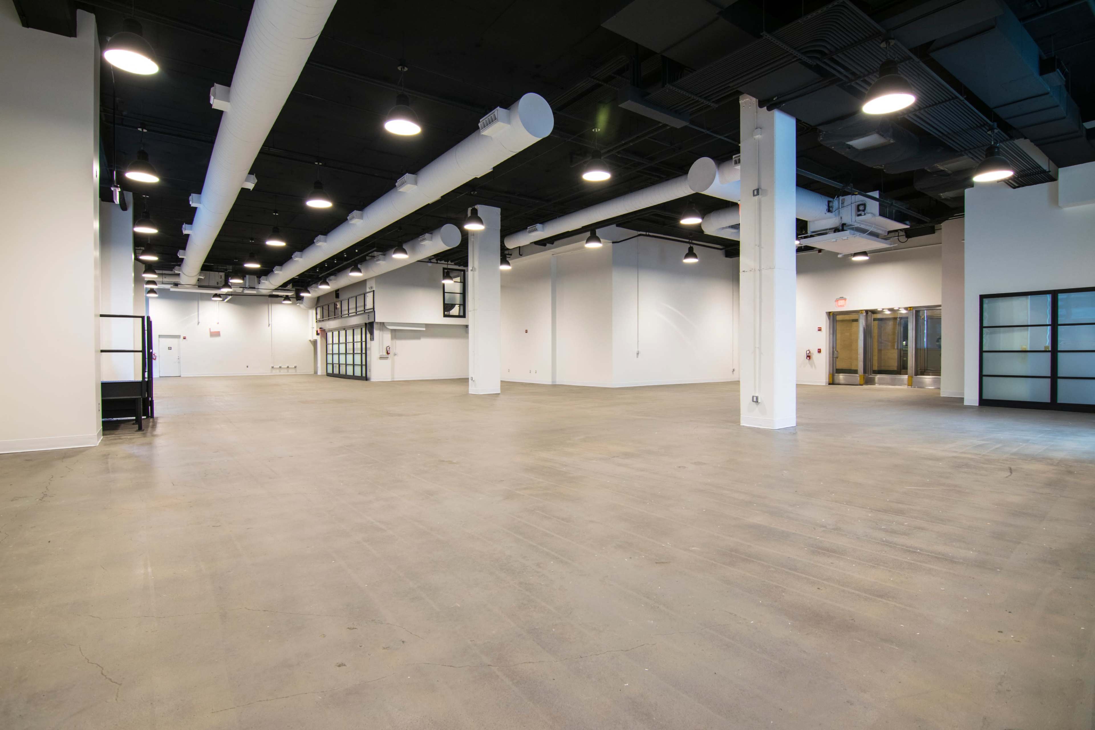 An empty industrial space with concrete floors and overhead pipes, featuring large windows and flat walls.