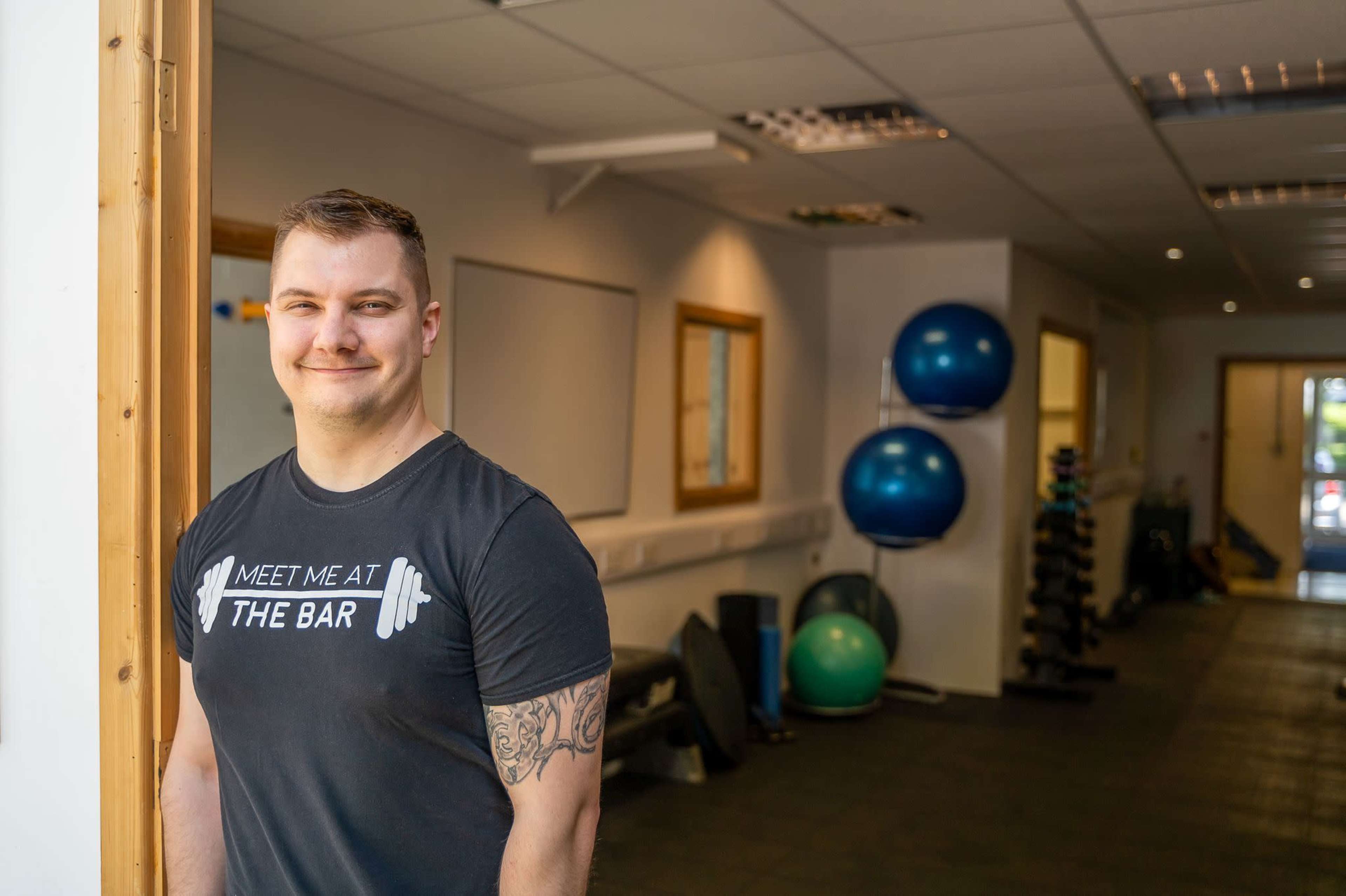 A man wearing a black t-shirt stands at the entrance of a gym, with fitness equipment and exercise balls visible in the background.