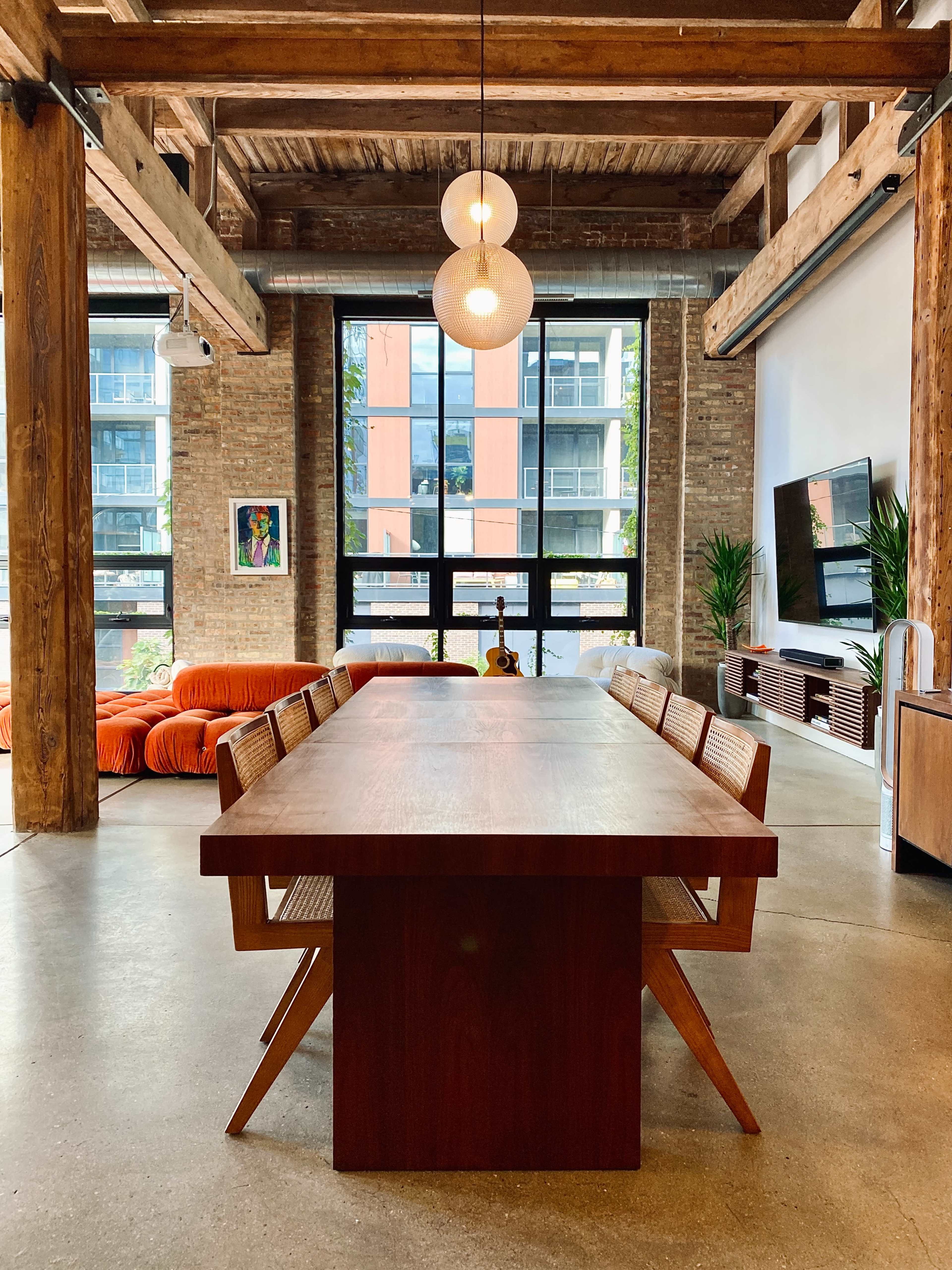 A modern dining area featuring a large wooden table surrounded by eight chairs, with exposed wooden beams and large windows in a bright, spacious room.