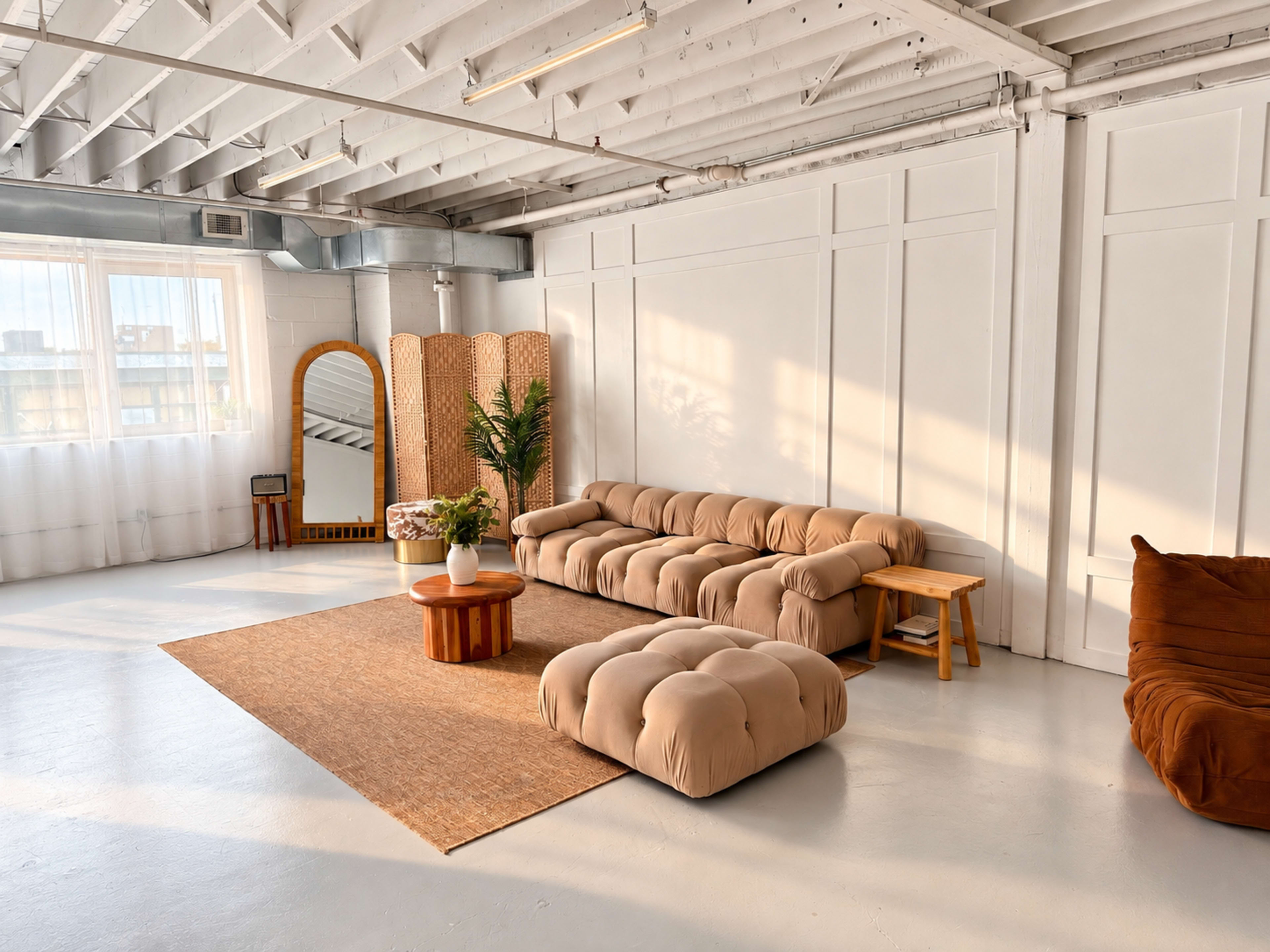 The image depicts a spacious room with white walls, hardwood floors, a large, light-colored sofa, a round wooden coffee table, and a decorative rug, all illuminated by natural light from large windows.