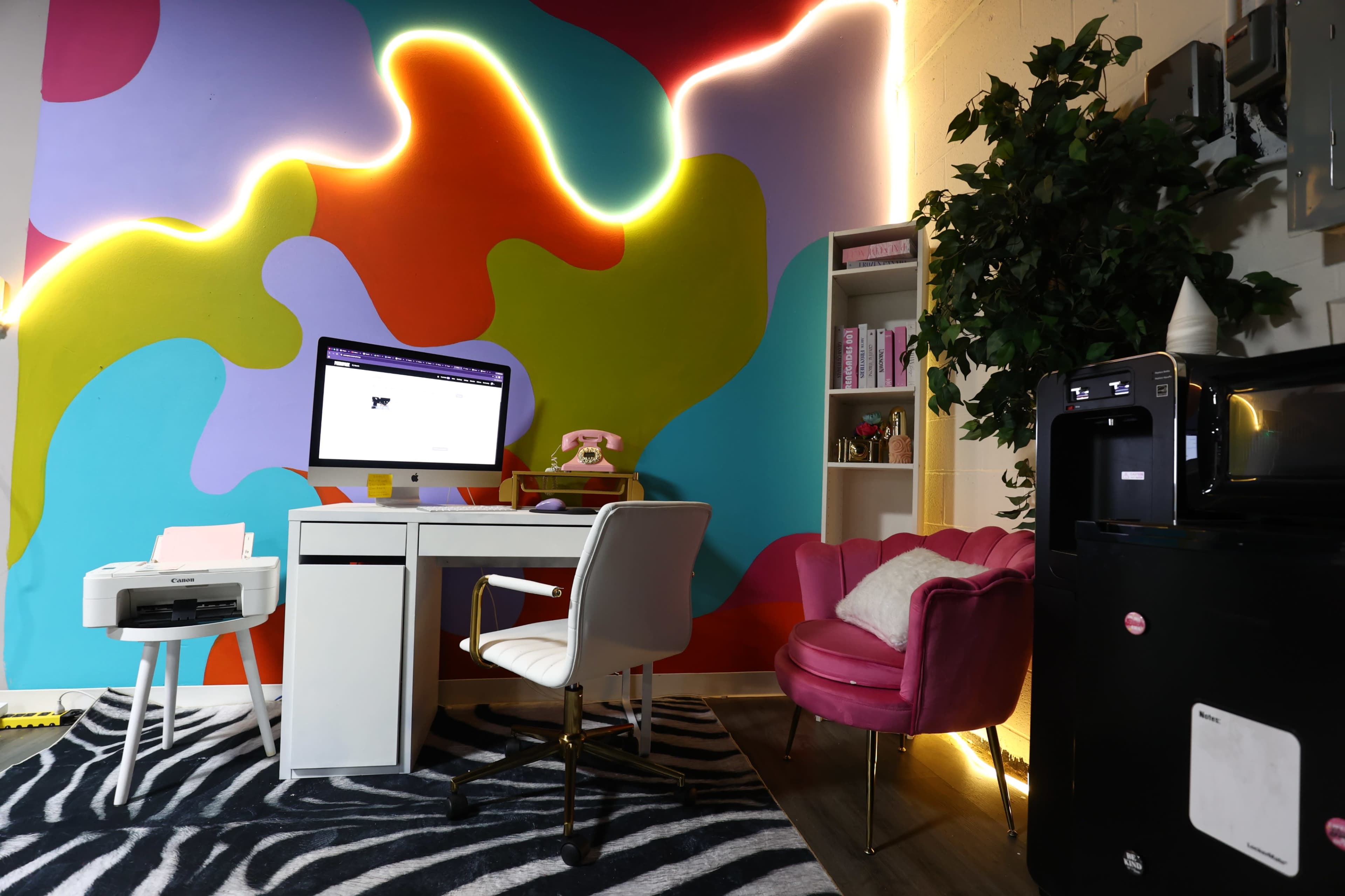The image shows a colorful home office with a white desk, an office chair, a computer monitor, a pink chair, a bookshelf, and appliances, all set against a vibrant abstract wall mural.