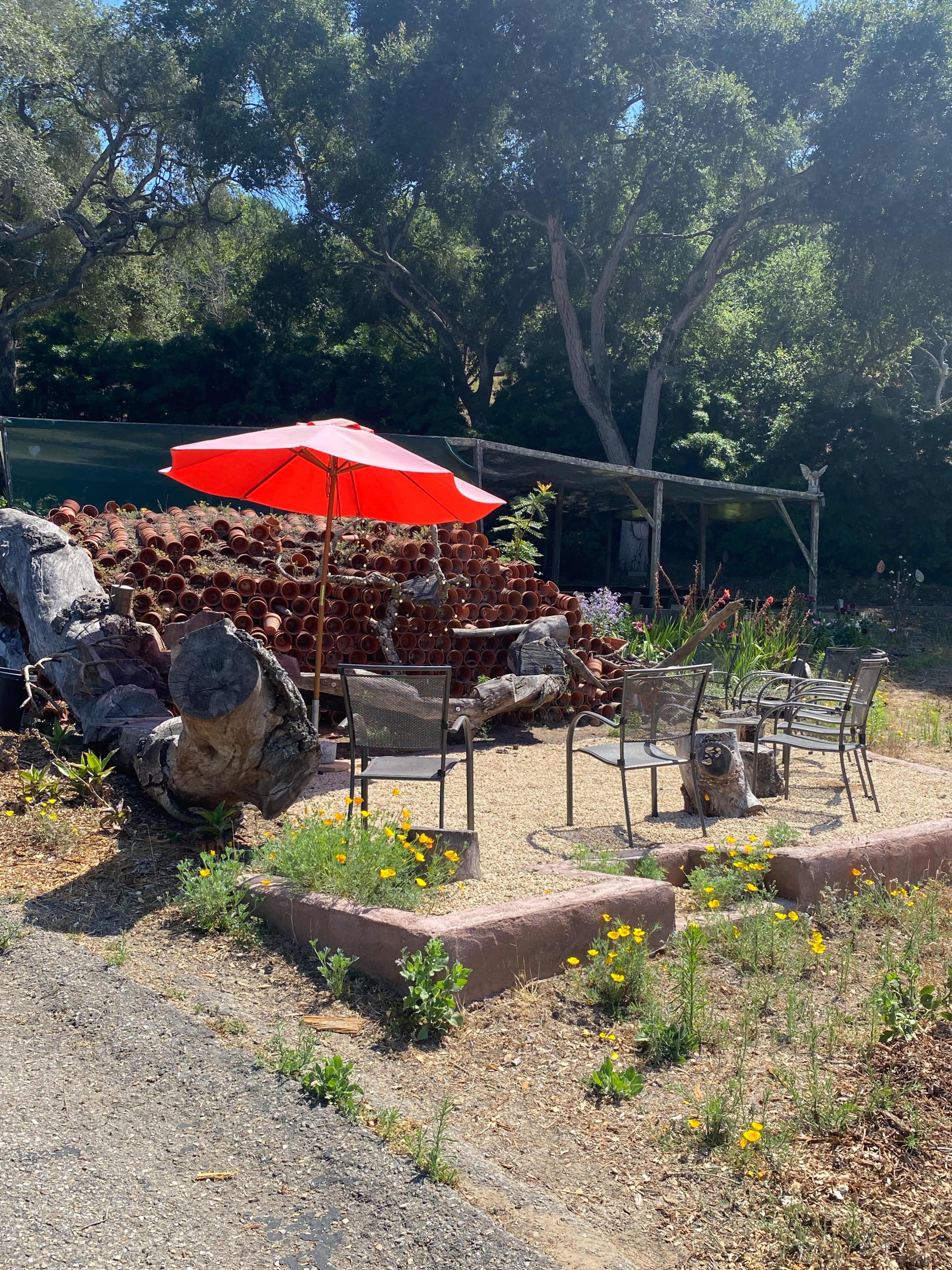 A red umbrella shades a set of chairs arranged around a gravel area, surrounded by potted plants and a rustic wooden feature.