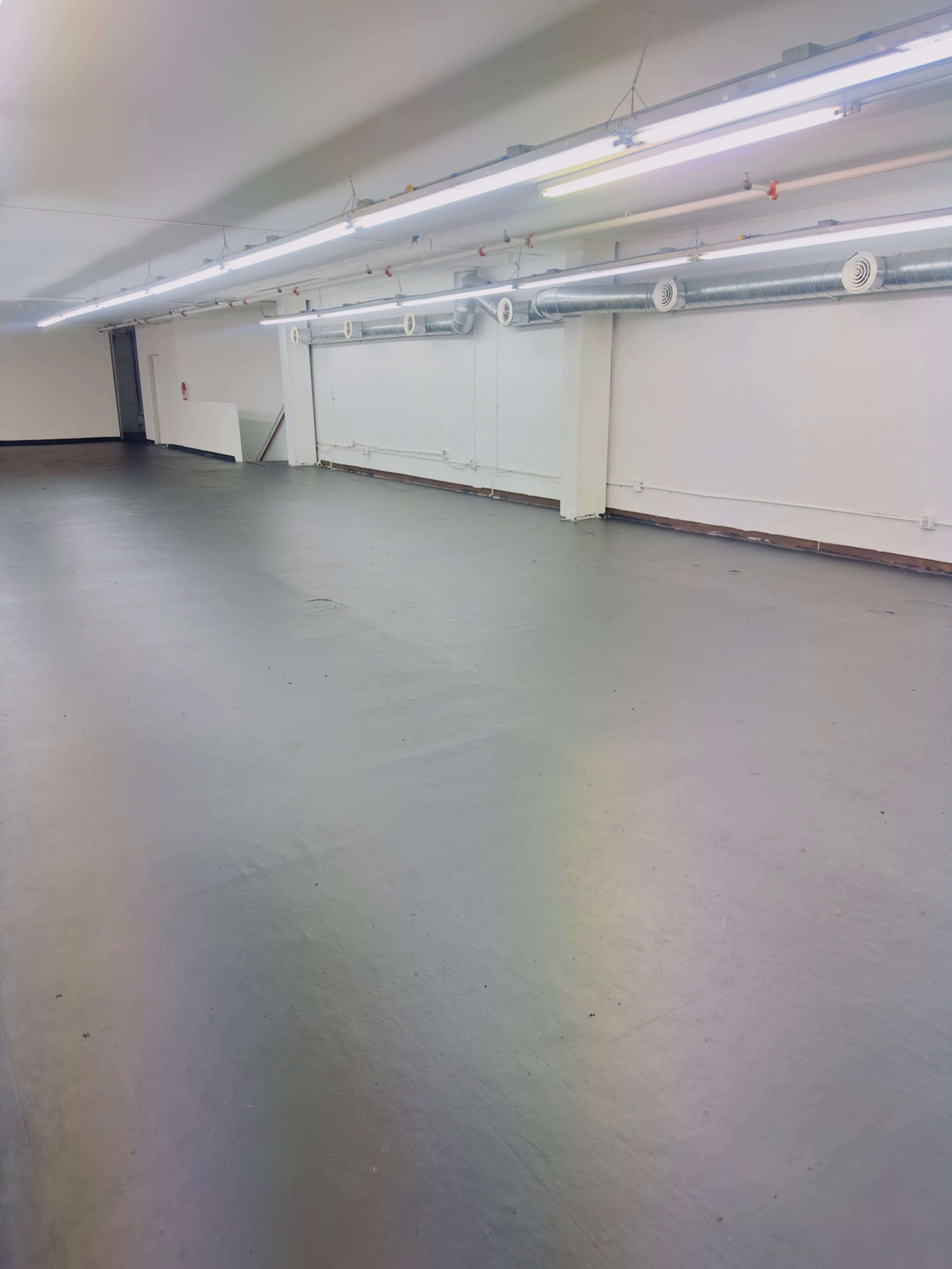 The image shows a spacious, empty room with a smooth gray floor and fluorescent lighting along the ceiling.