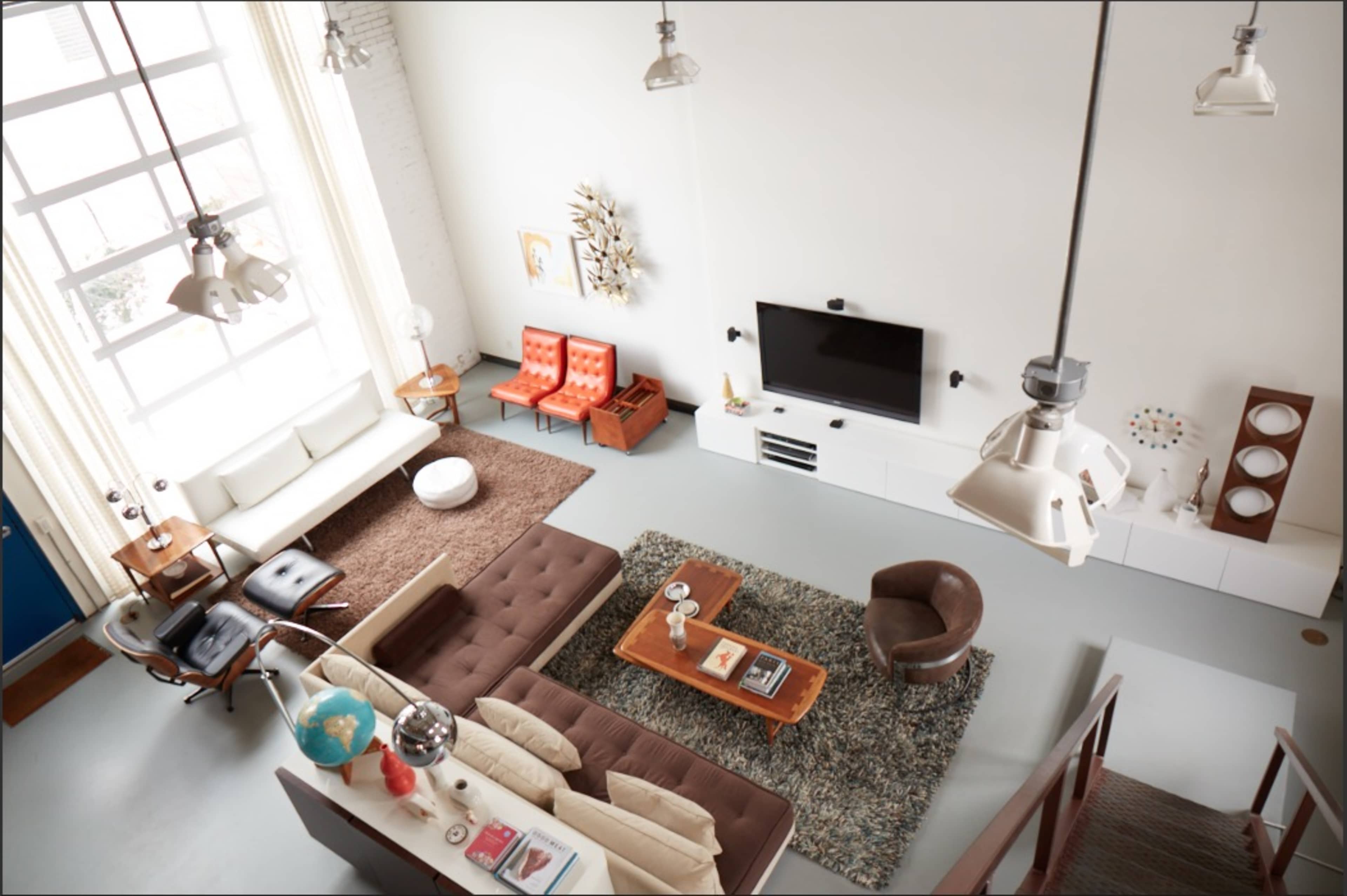 The image depicts a modern living room with a large window, featuring two orange chairs, a brown sofa, a coffee table, and a television mounted on the wall.