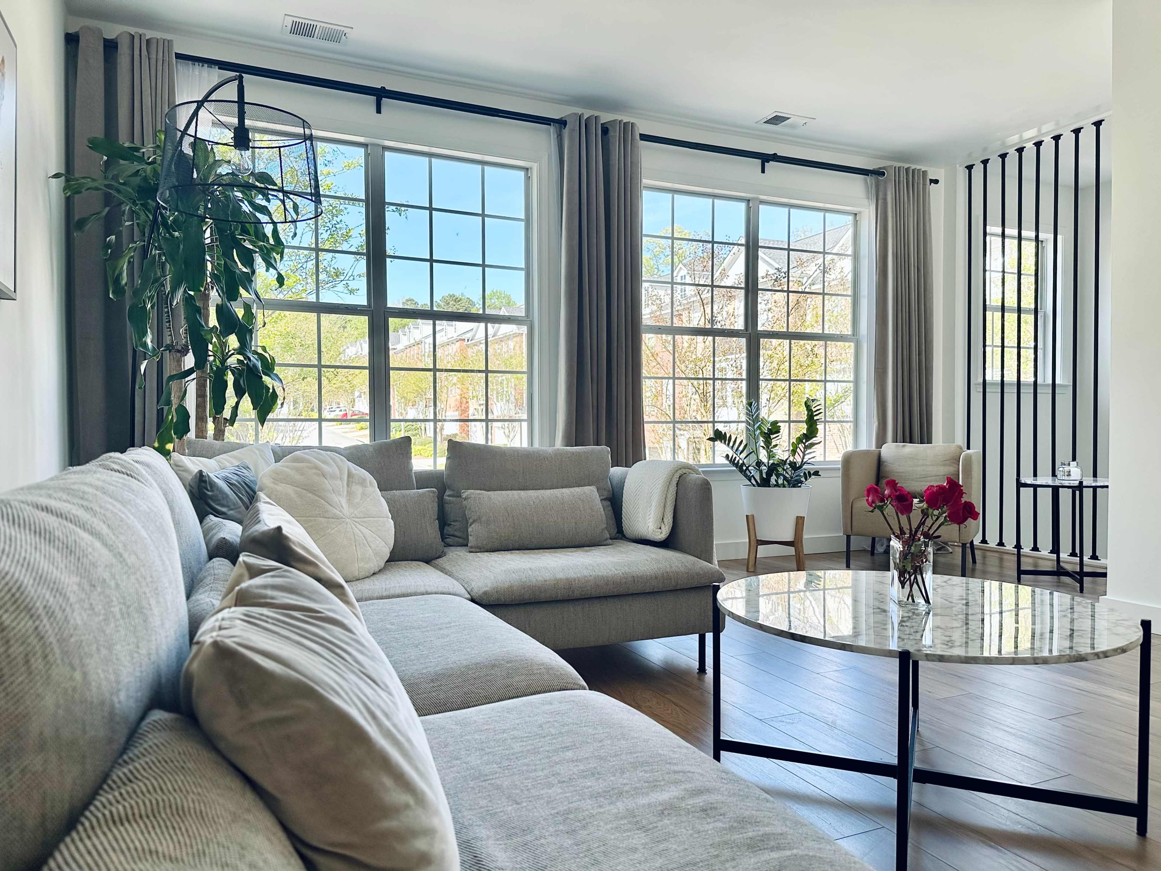 The image shows a bright living room with a sectional sofa, a round coffee table, large windows, and potted plants.