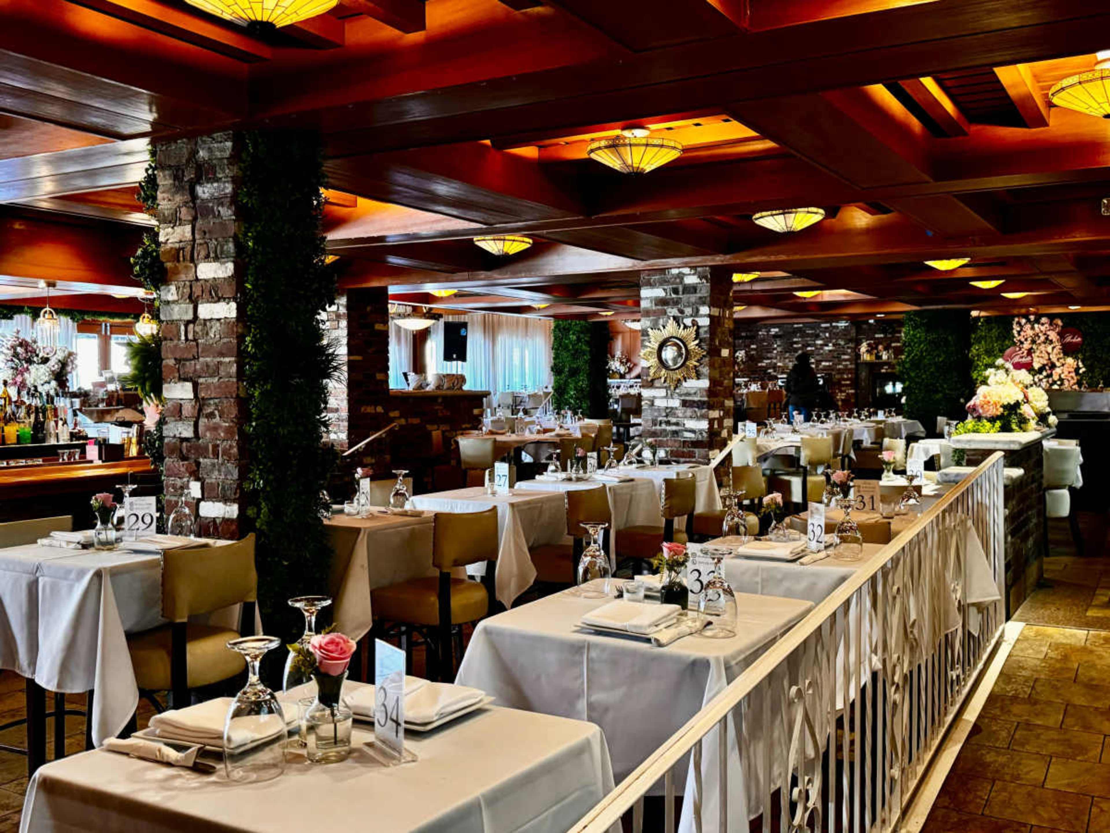 The image shows a spacious restaurant interior with neatly arranged tables, each set with white tablecloths and glassware, surrounded by brick and wooden columns.