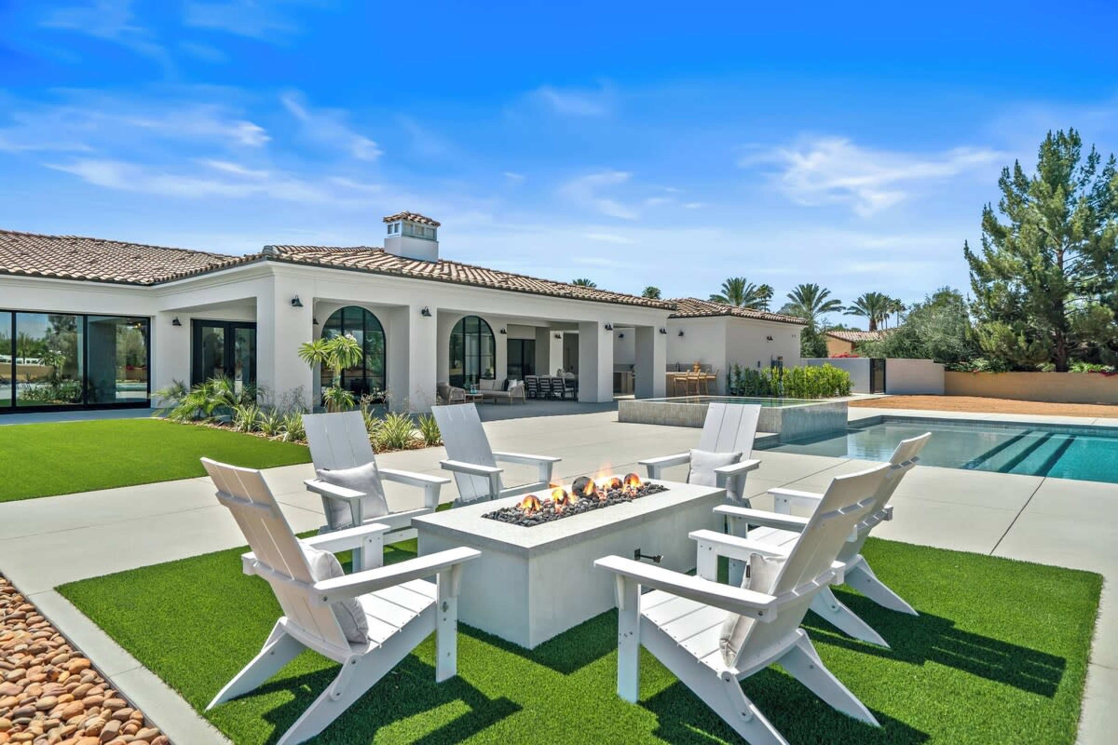 A modern backyard features a fire pit surrounded by white chairs, a swimming pool, and a spacious house with large windows and lush landscaping.