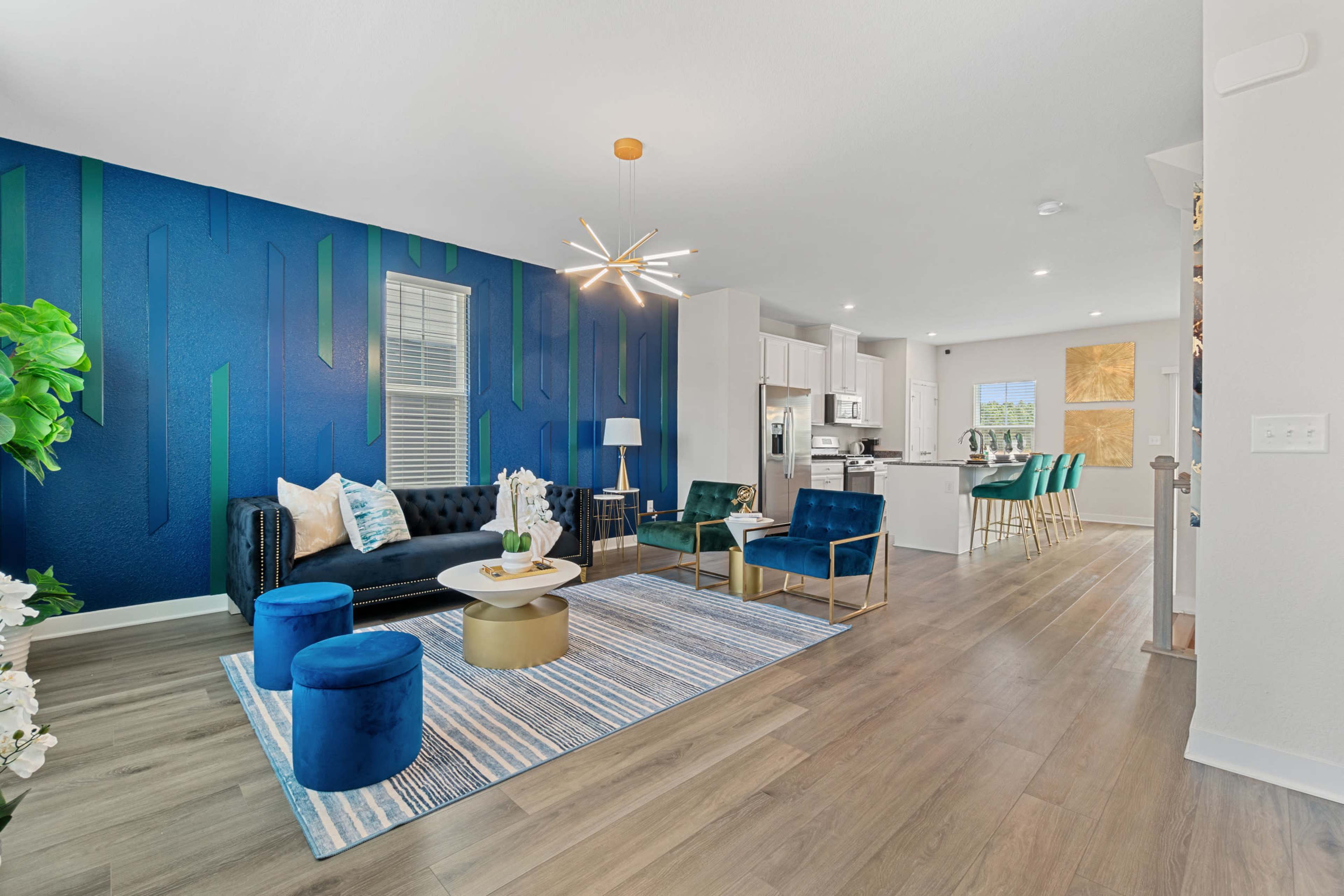 A modern living room with a navy blue accent wall, a black sofa, and teal seating, complemented by a light wood floor and a kitchen visible in the background.