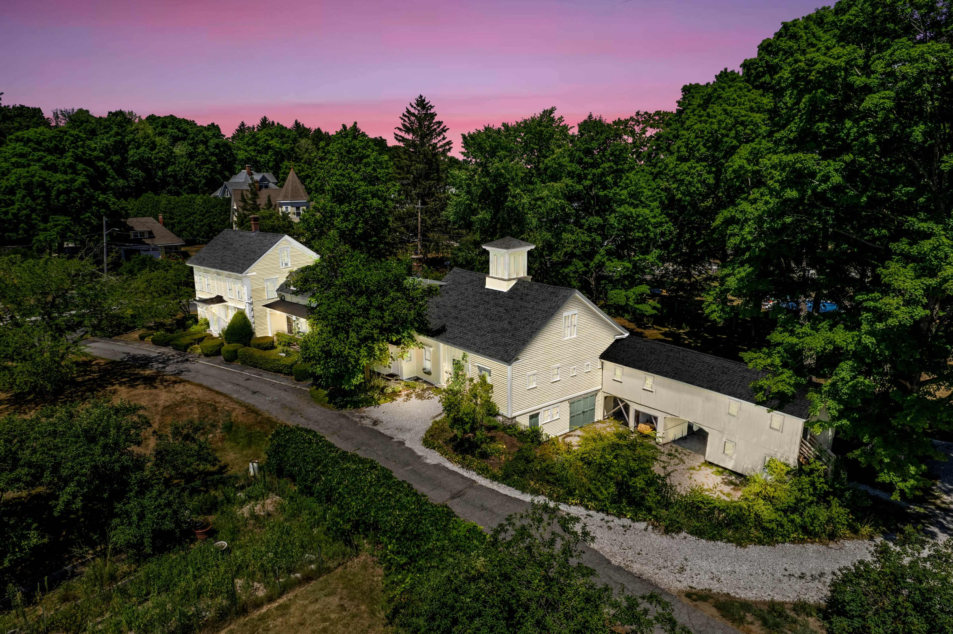 A large, white, two-story building with a bell tower is surrounded by lush greenery and a gravel driveway.