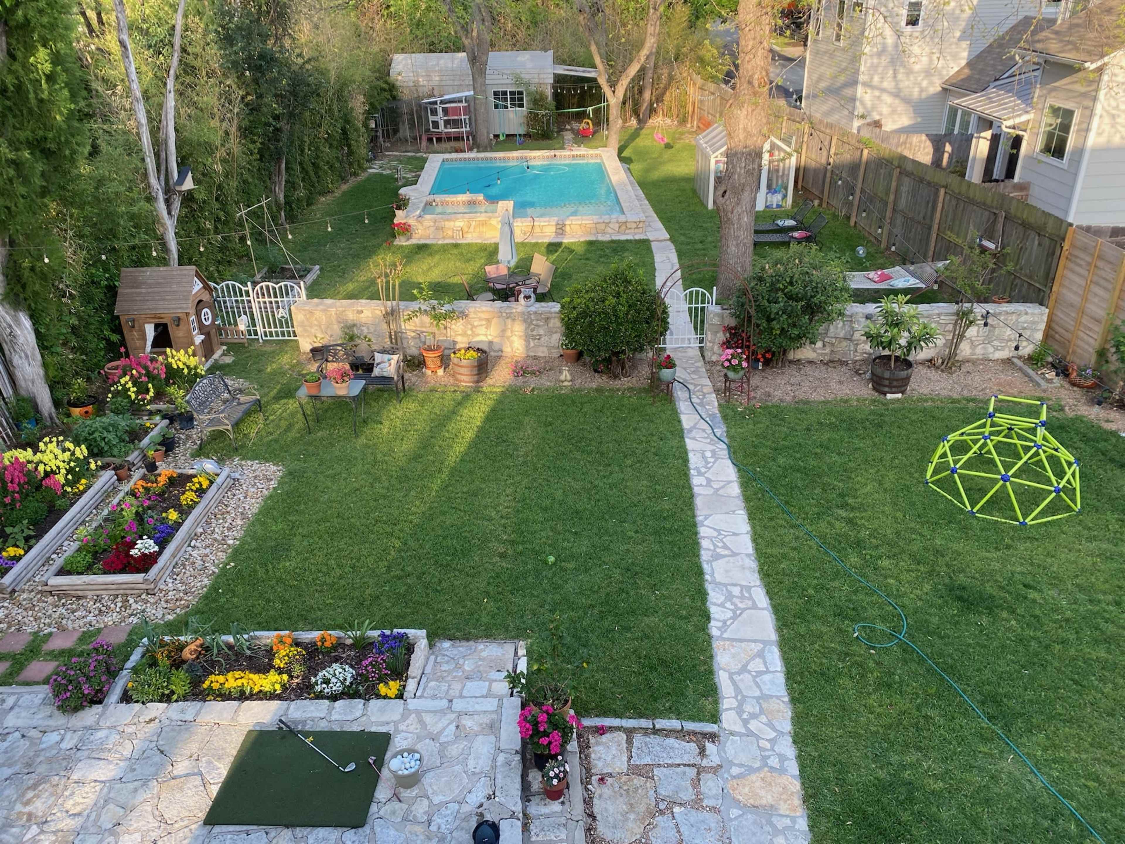 A landscaped backyard featuring a swimming pool, garden beds with colorful flowers, a stone path, and a children's play structure.