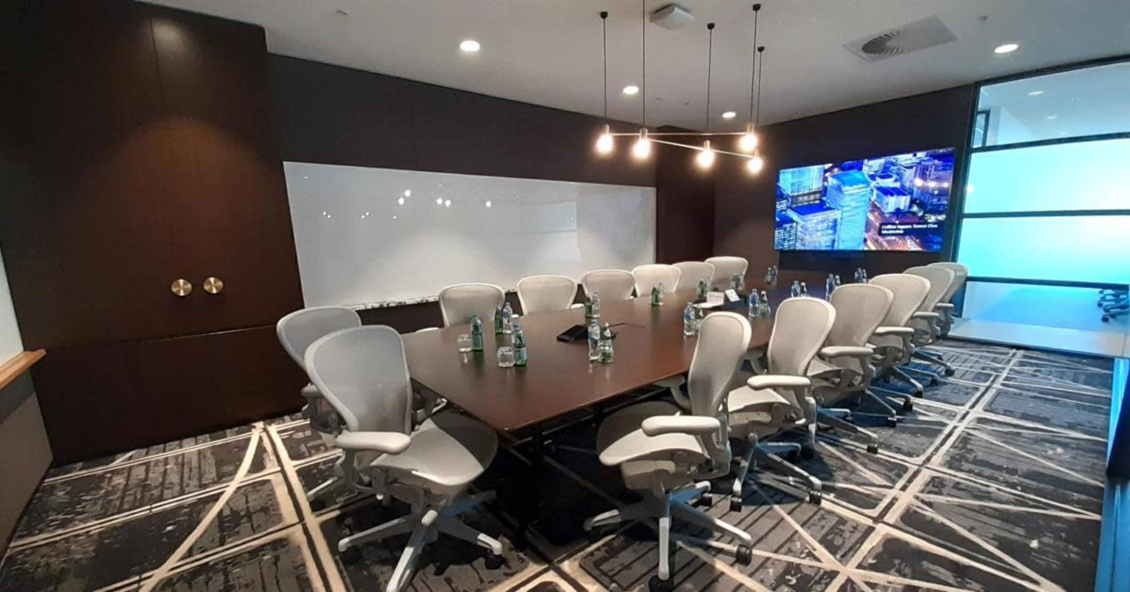 A large conference room features a long table surrounded by ergonomic chairs, a whiteboard, and a screen displaying an image.