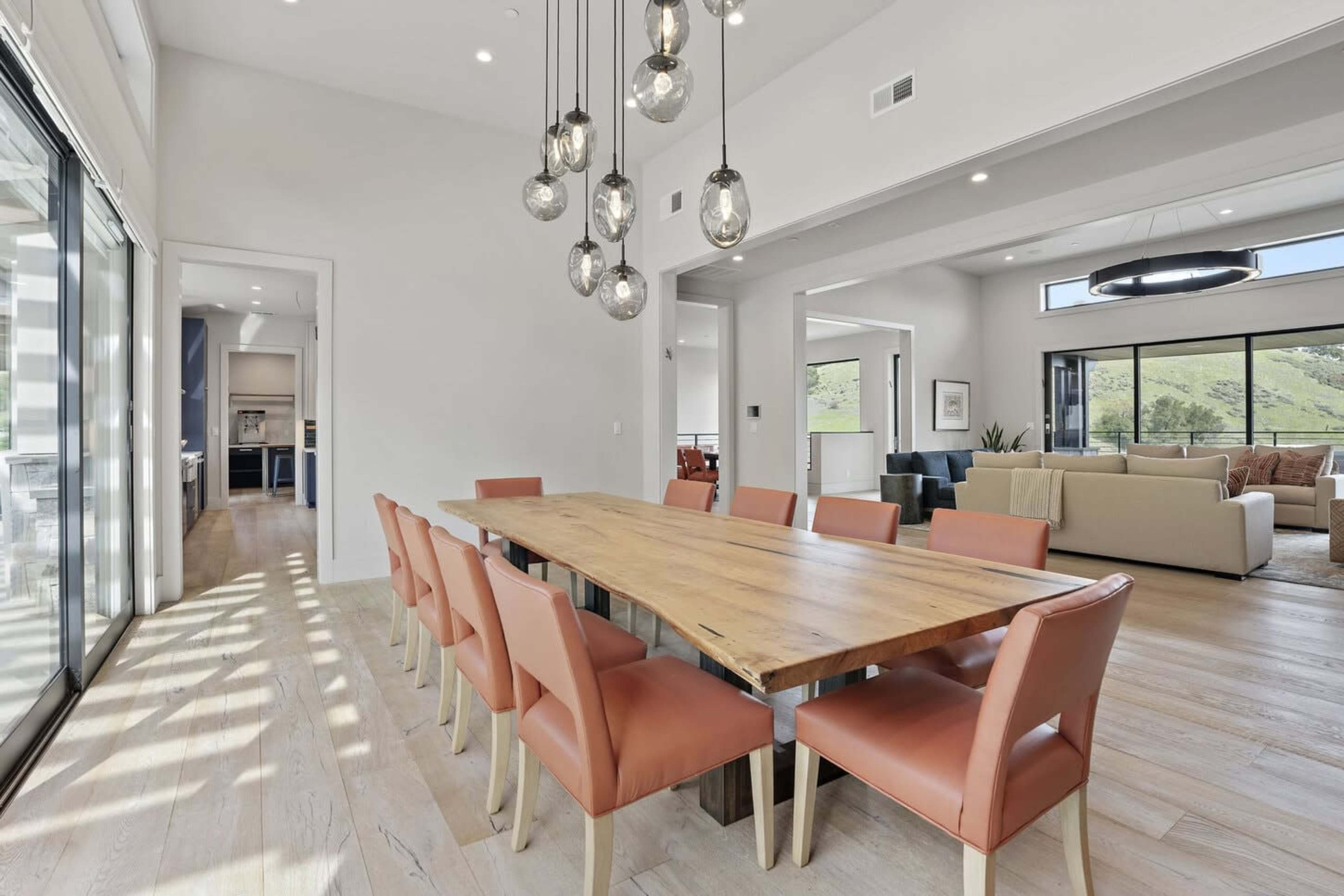 The image shows a modern dining area with a long wooden table surrounded by eight orange chairs, illuminated by hanging pendant lights.