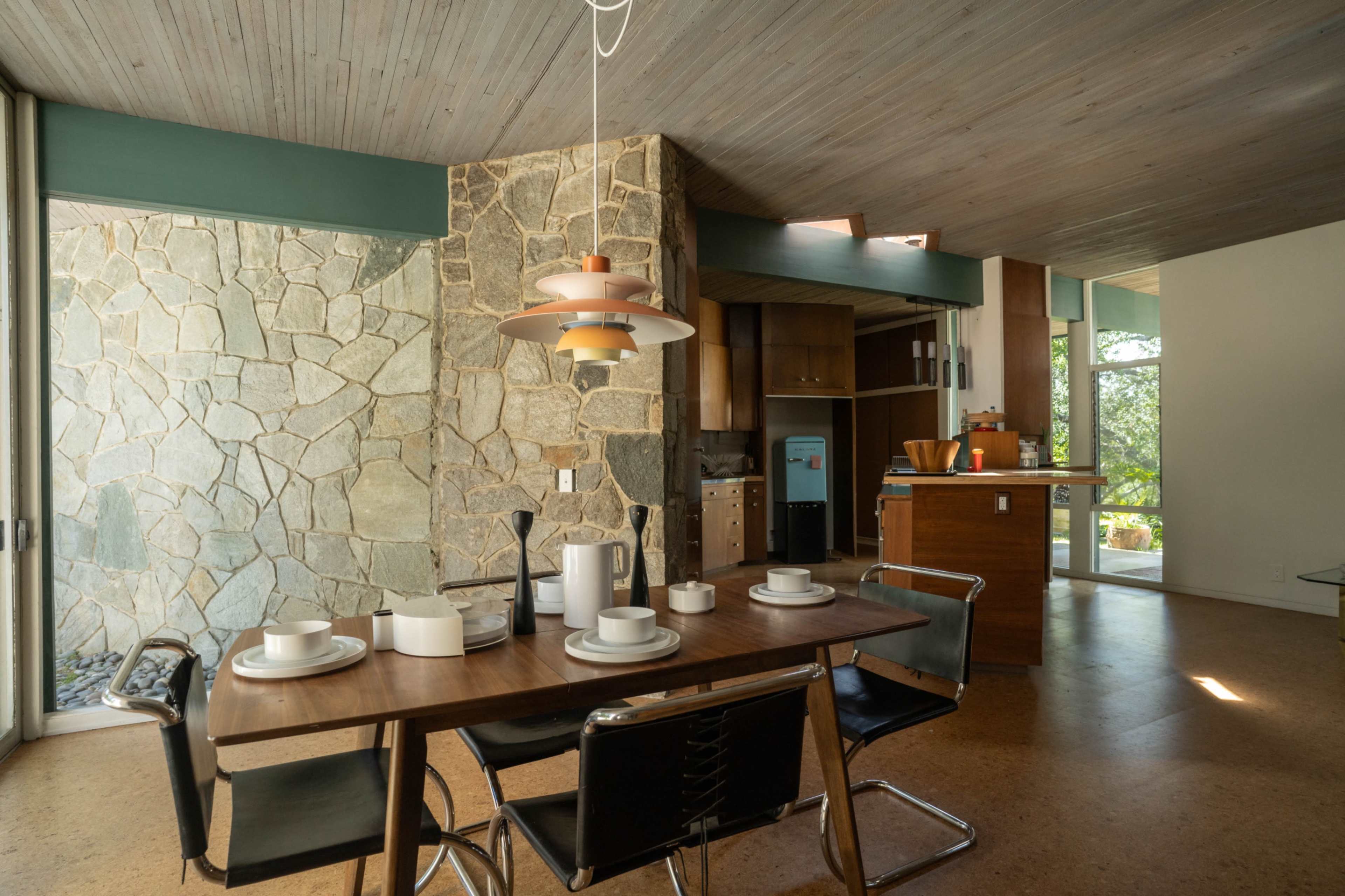 The image shows a modern kitchen and dining area featuring a wooden table set for a meal, a stone wall, and large windows that allow natural light to enter.