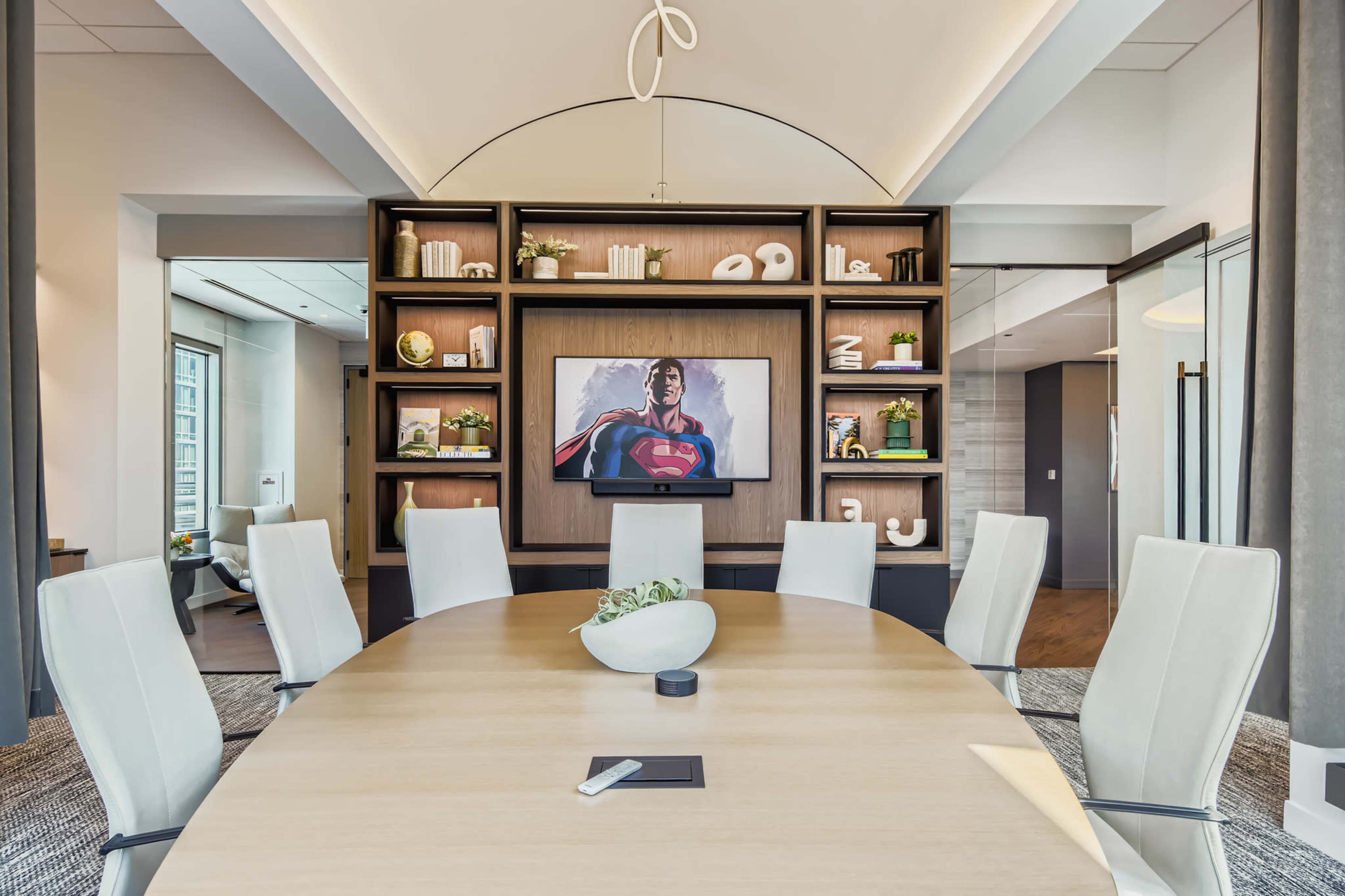 A modern conference room features a large oval table surrounded by white chairs, with a wall shelf displaying decorative items and a framed image of a superhero.