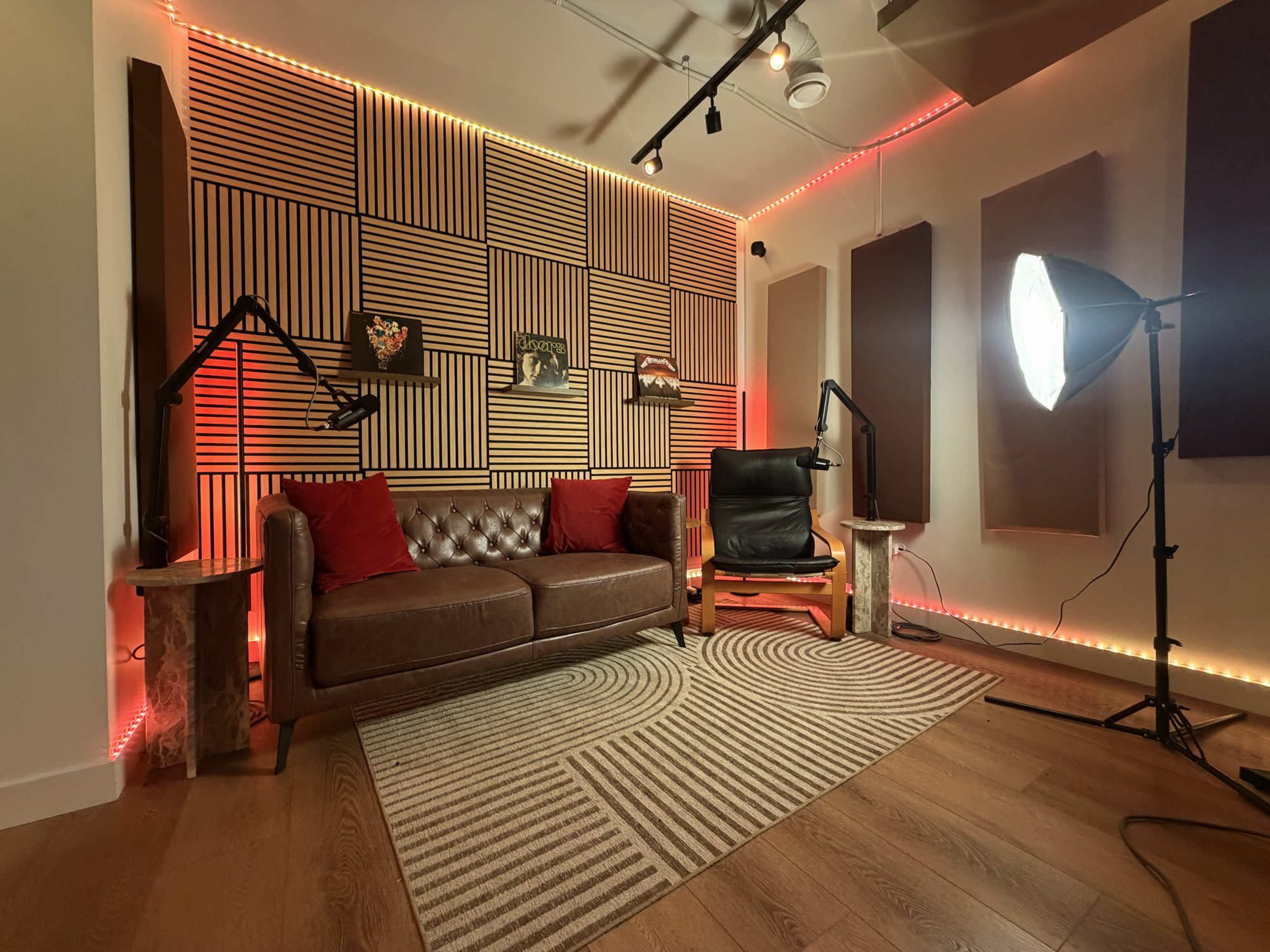 The image shows a recording or podcast studio featuring a leather sofa, a comfortable chair, and sound-absorbing wall panels, illuminated by colored LED lights.