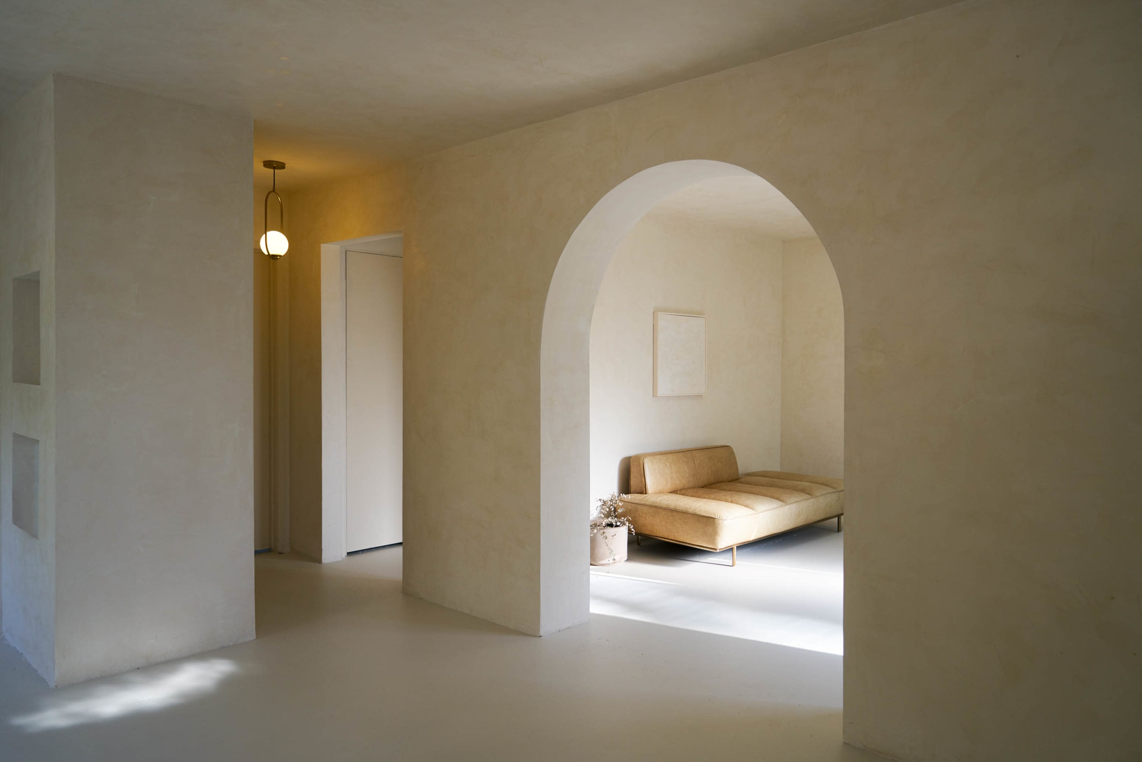 A minimalistic interior space featuring a light-colored archway, a beige sofa, and soft natural lighting.