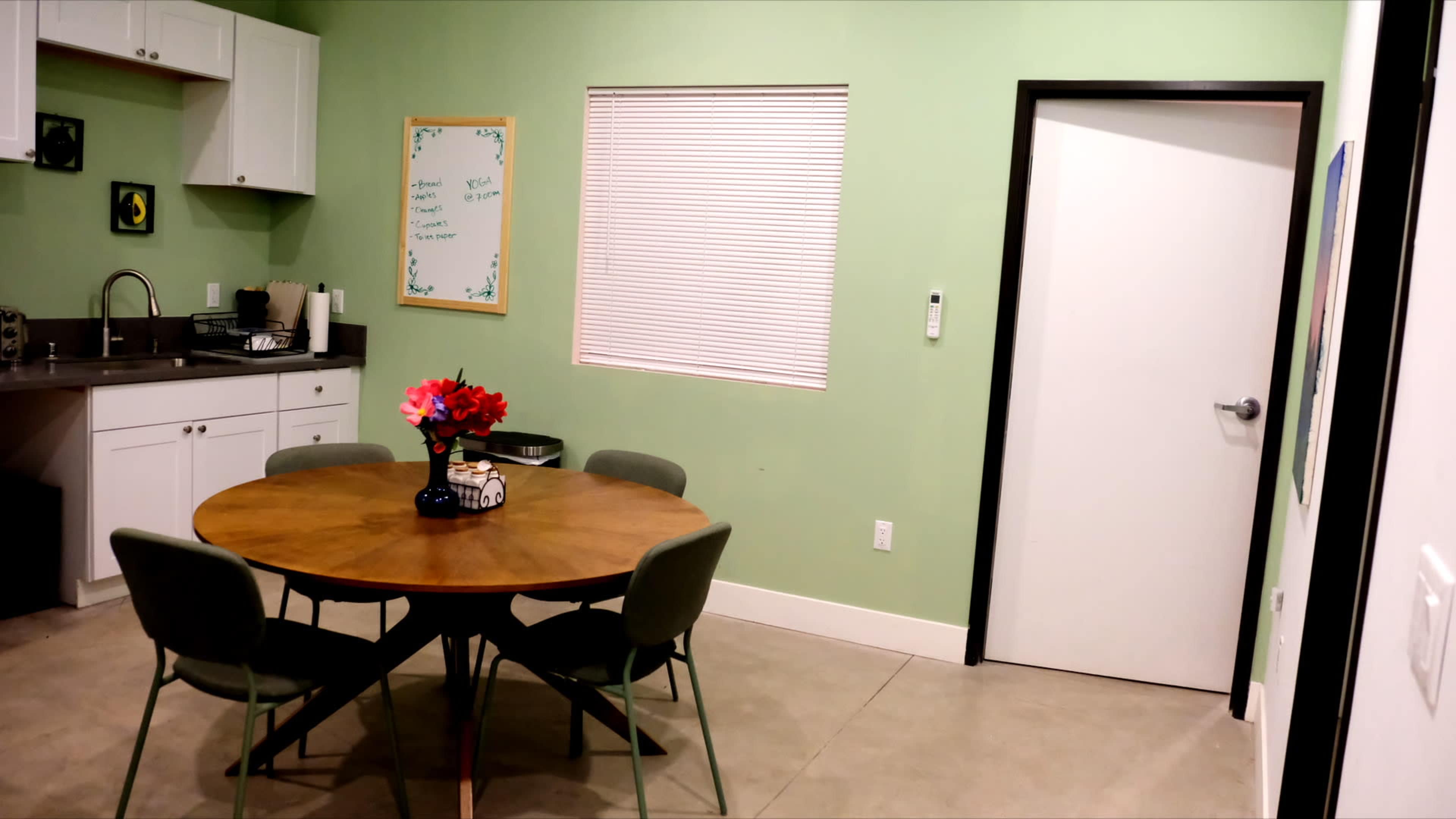 The image shows a simple dining area with a round wooden table surrounded by four green chairs, along with a kitchenette and a door leading to another room.