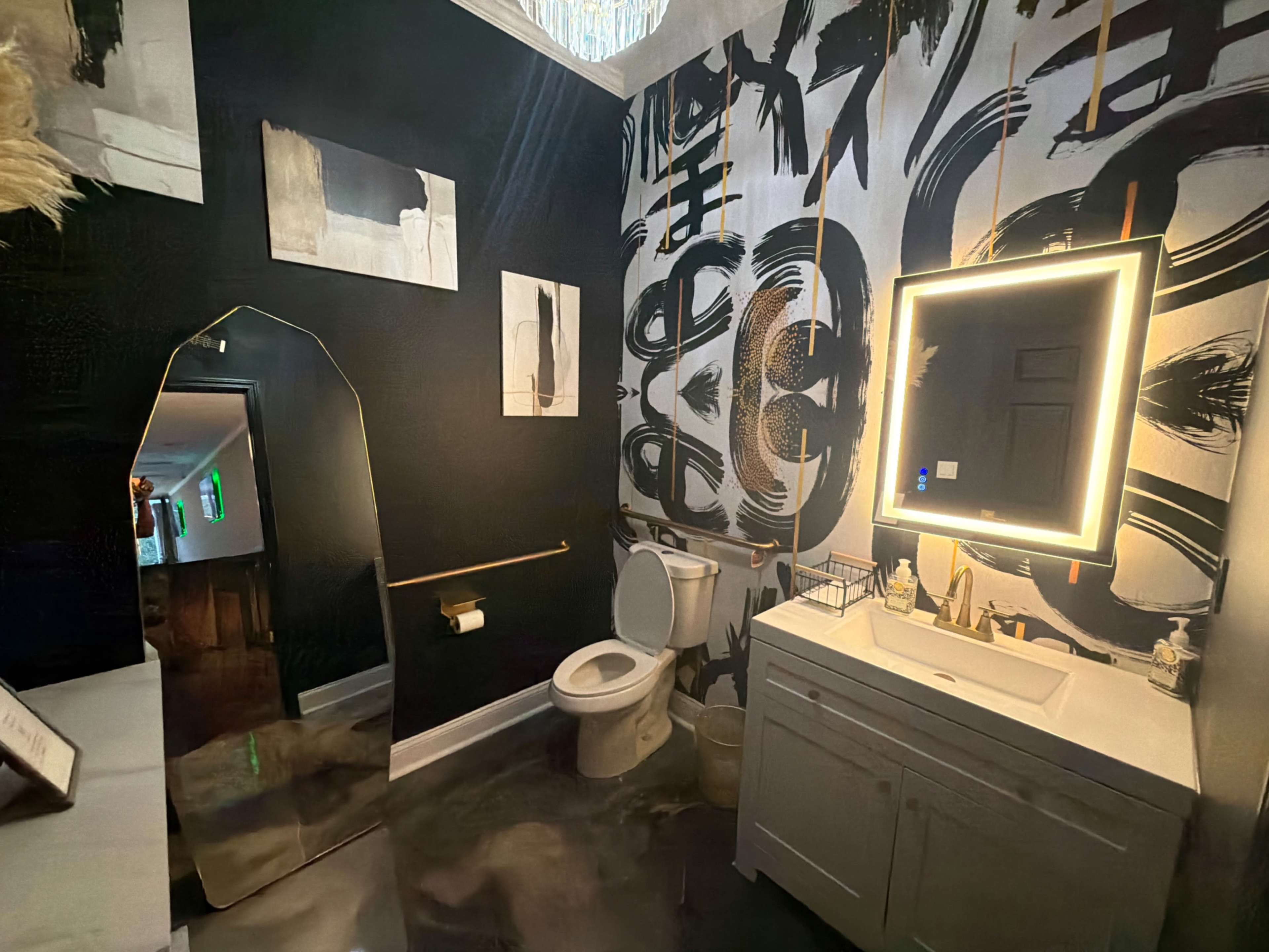 The image depicts a modern bathroom featuring black walls adorned with abstract artwork, a white toilet, a sleek vanity with an illuminated mirror, and a large decorative mirror.