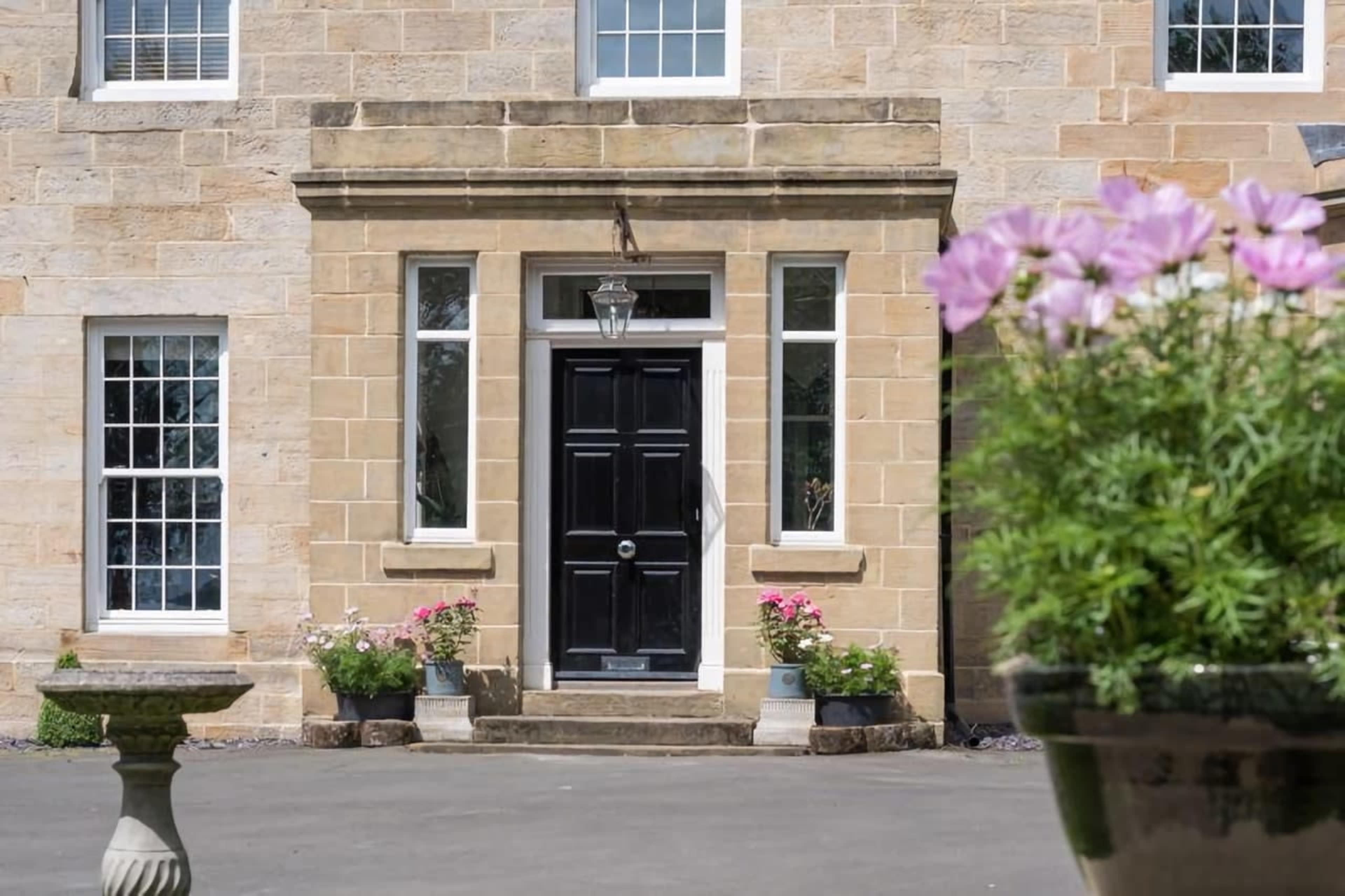 The image shows a stone facade of a house with a black front door framed by tall windows and flower pots on either side.