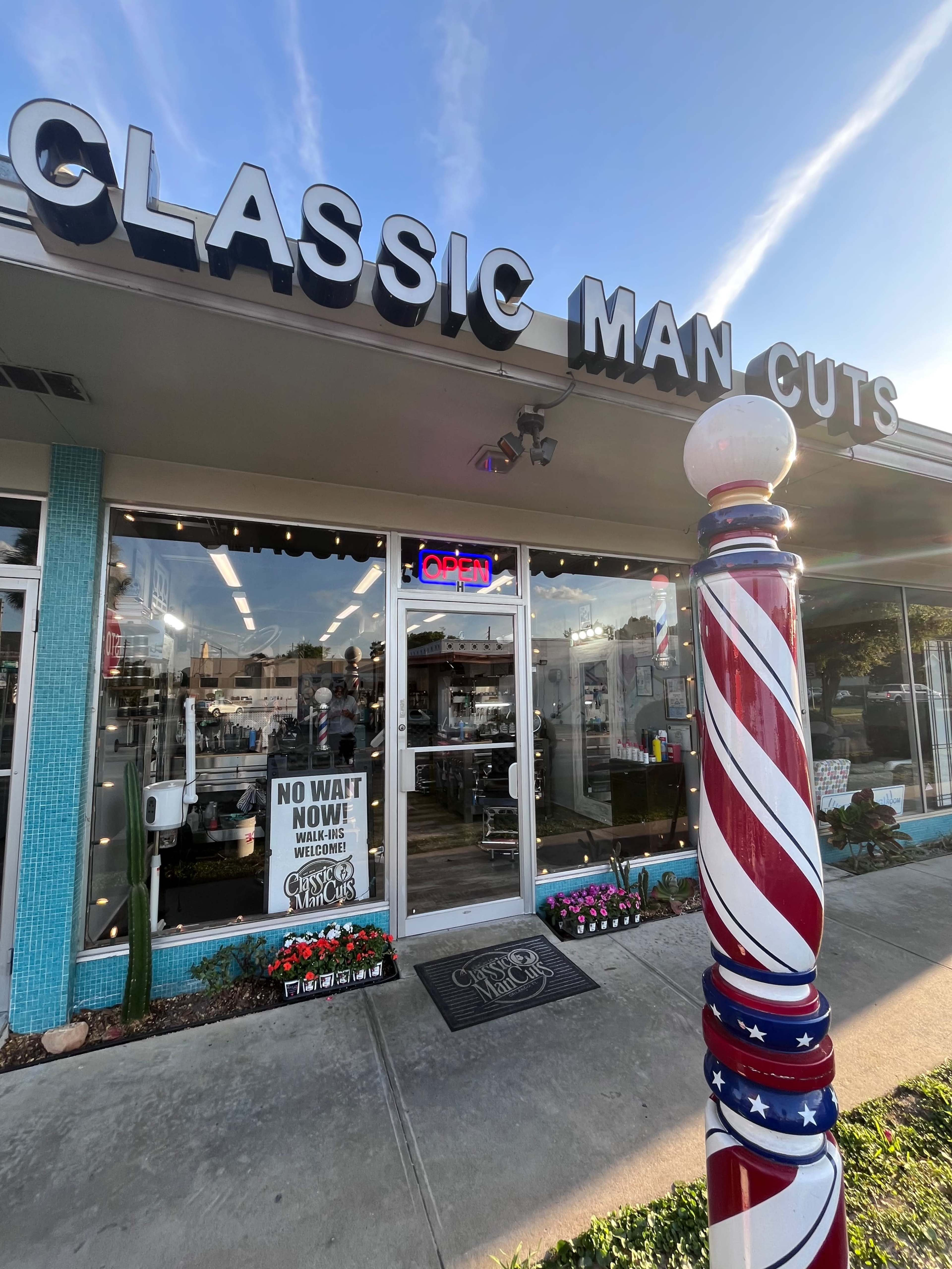 The front of a barbershop named "Classic Man Cuts" features a brightly lit sign, an open door, and a red, white, and blue barber pole.