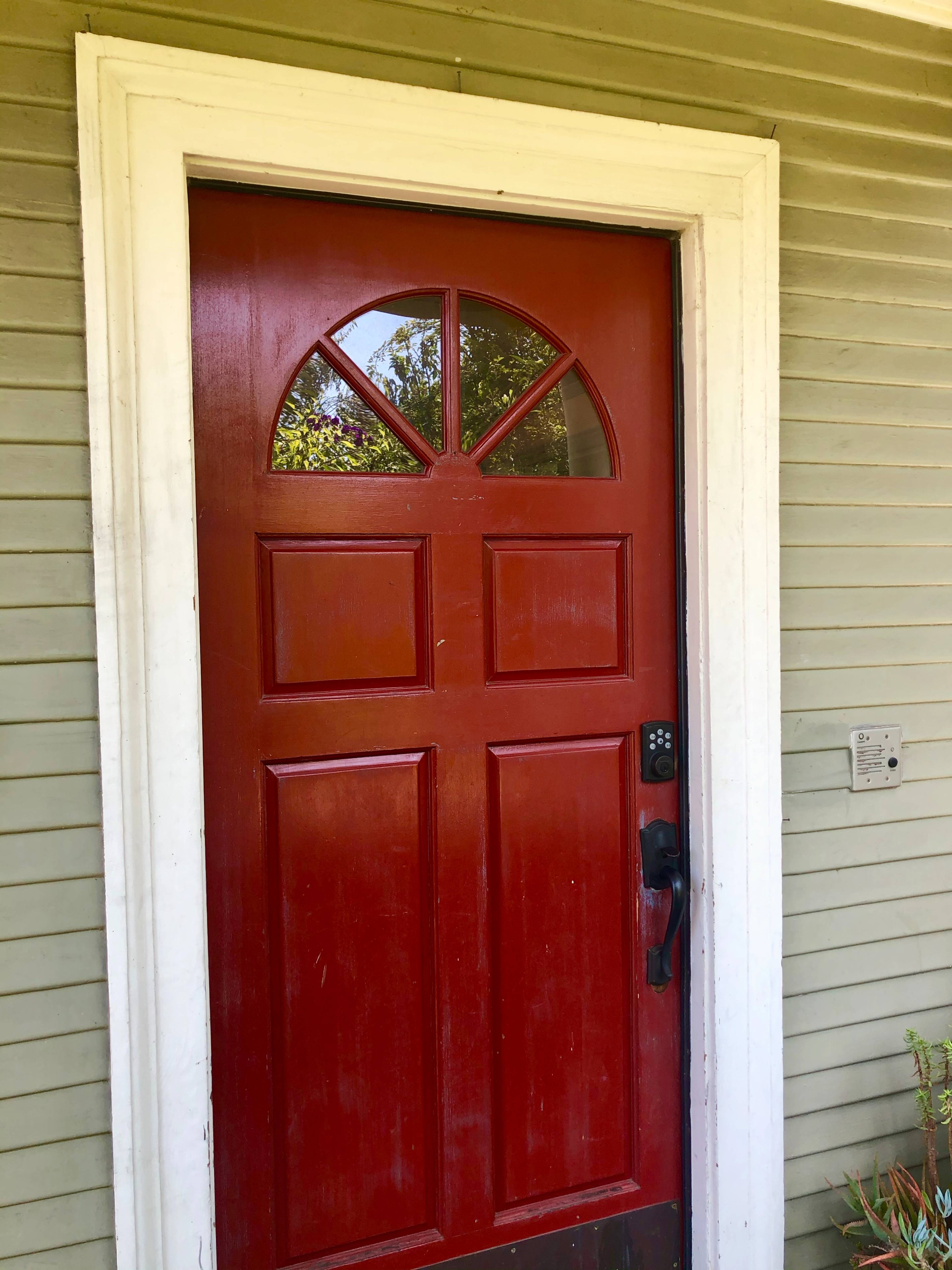 A red front door with a decorative upper panel and a black door handle, situated on a light green house exterior.