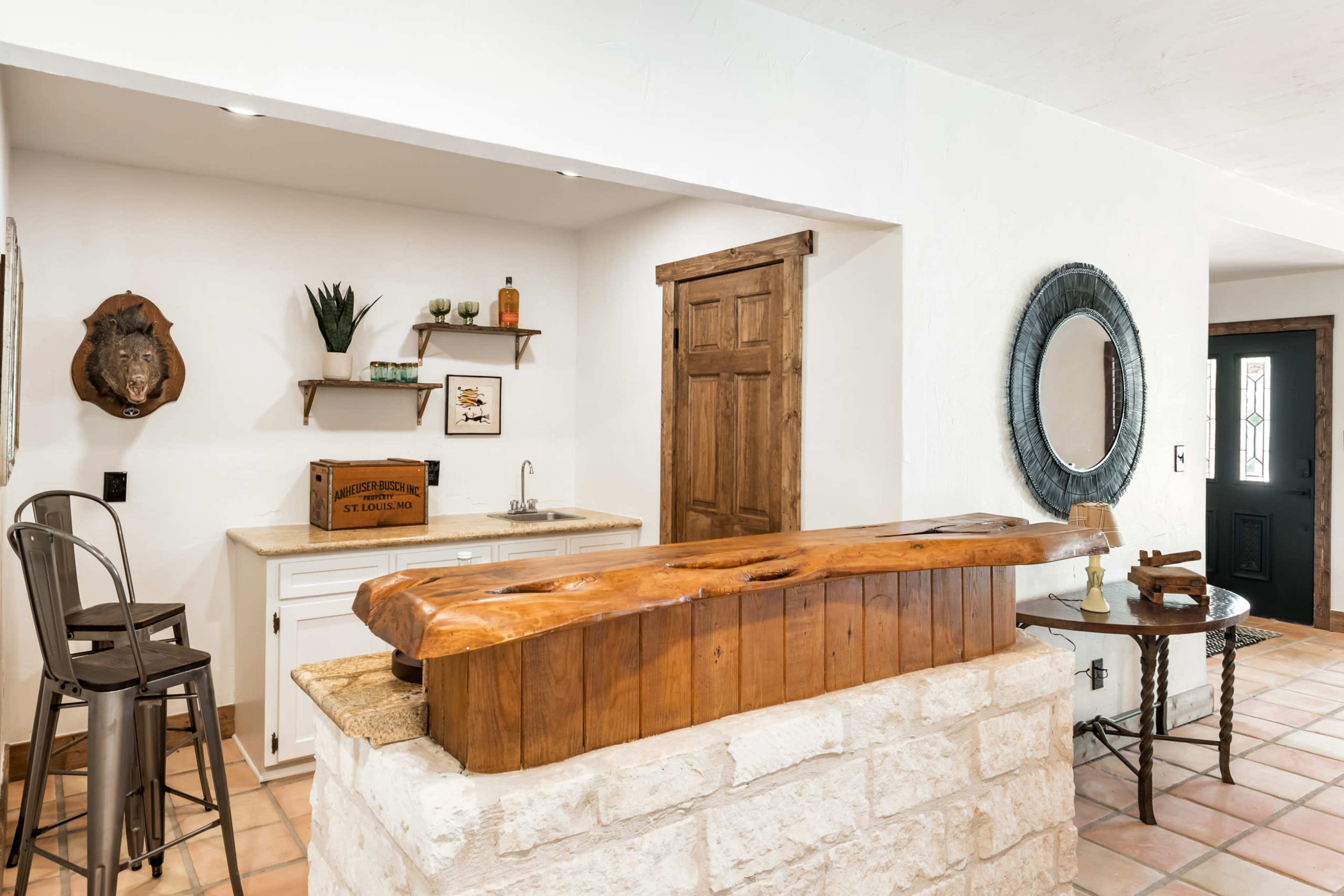 A rustic kitchen with a wooden bar counter, a sink, and a wooden door, complemented by stone and tiled flooring.