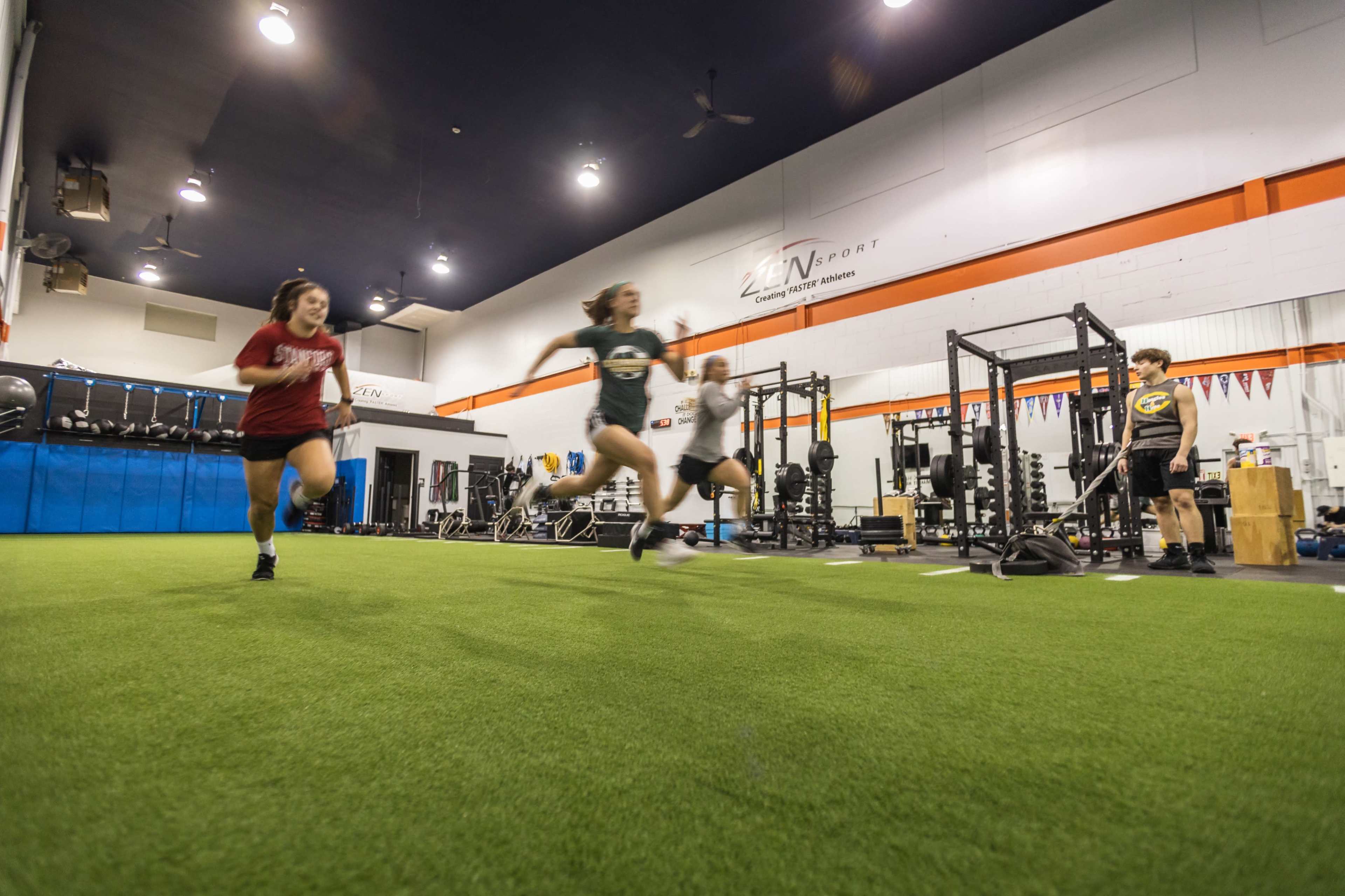 People are running on a turf surface inside a training facility equipped with weightlifting racks and exercise equipment.