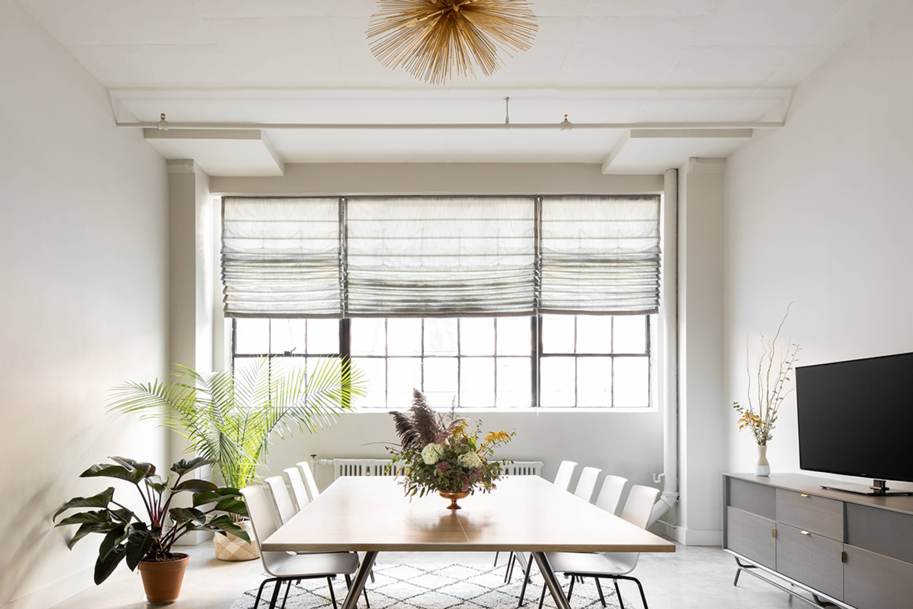 A spacious dining area features a large table surrounded by chairs, with a plant and a decorative arrangement on the table, and large windows with light-filtering shades.