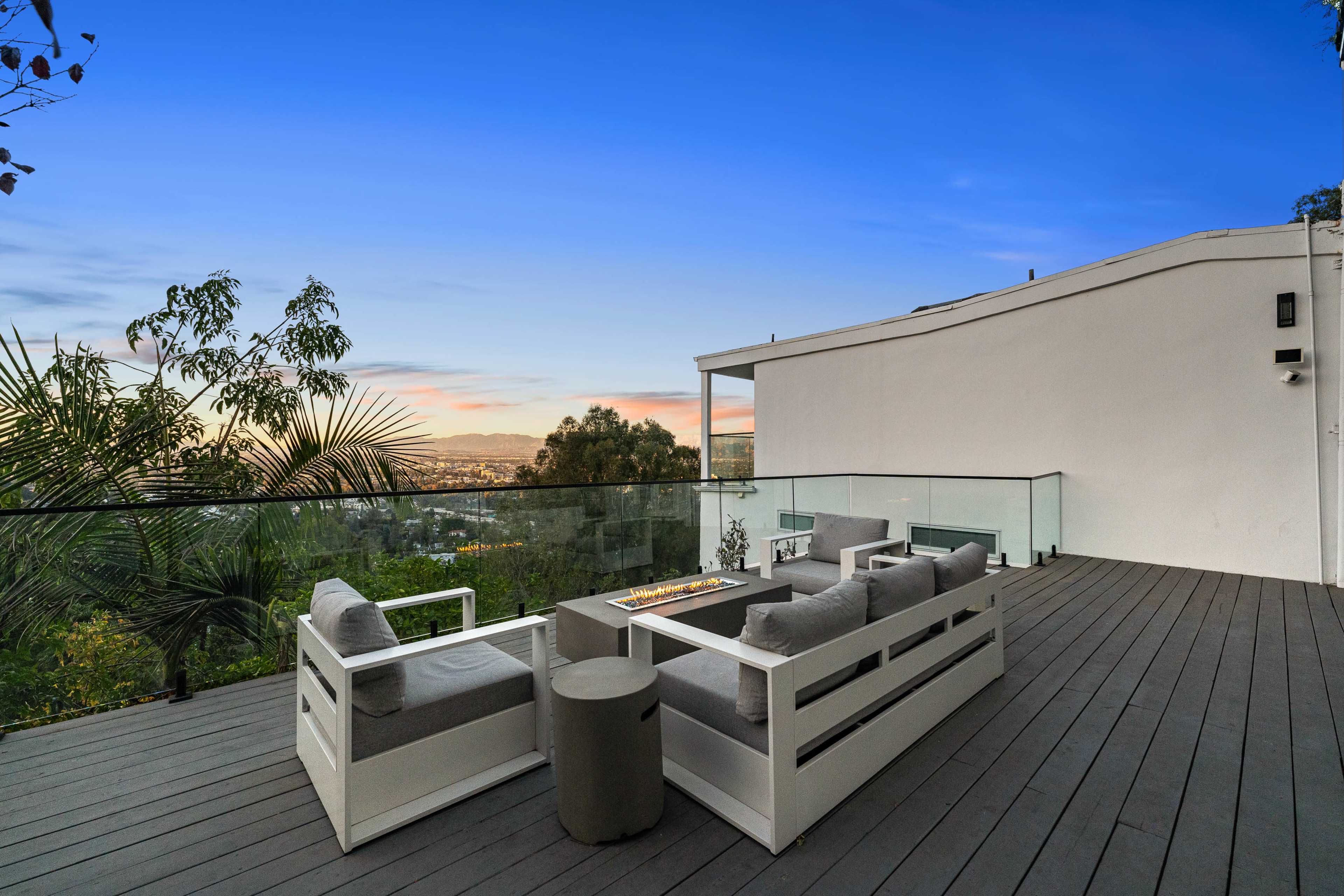 A modern outdoor terrace features a seating area with a fire pit, surrounded by a clear railing and overlooking a scenic view at sunset.