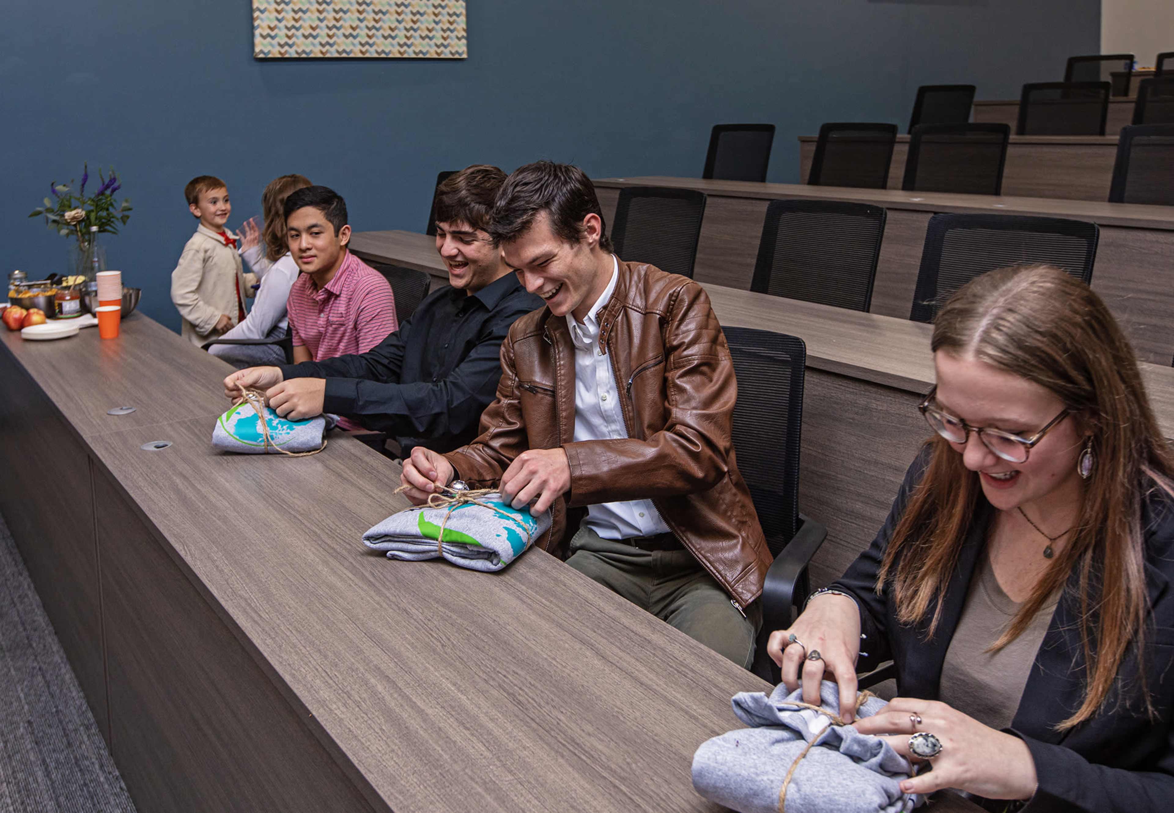 A group of six people sits at a long table, wrapping clothing items in a conference room.