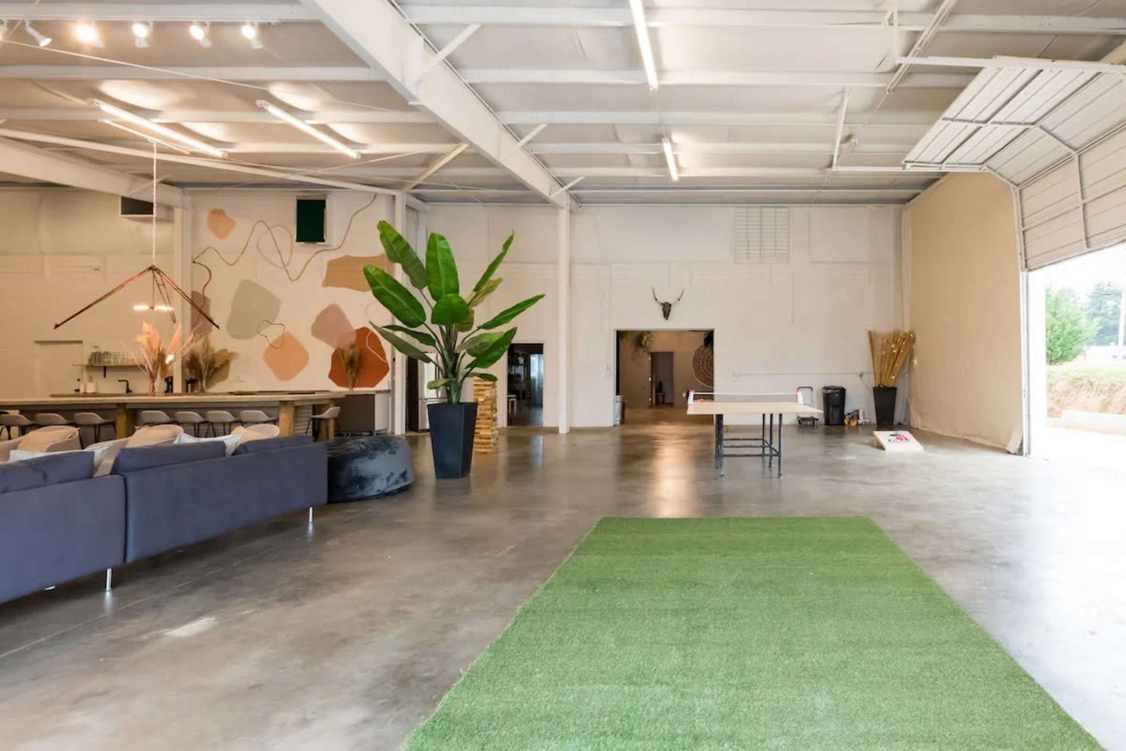 The image shows a spacious, modern indoor venue with a concrete floor, a large plant in a pot, and an area covered in green turf.