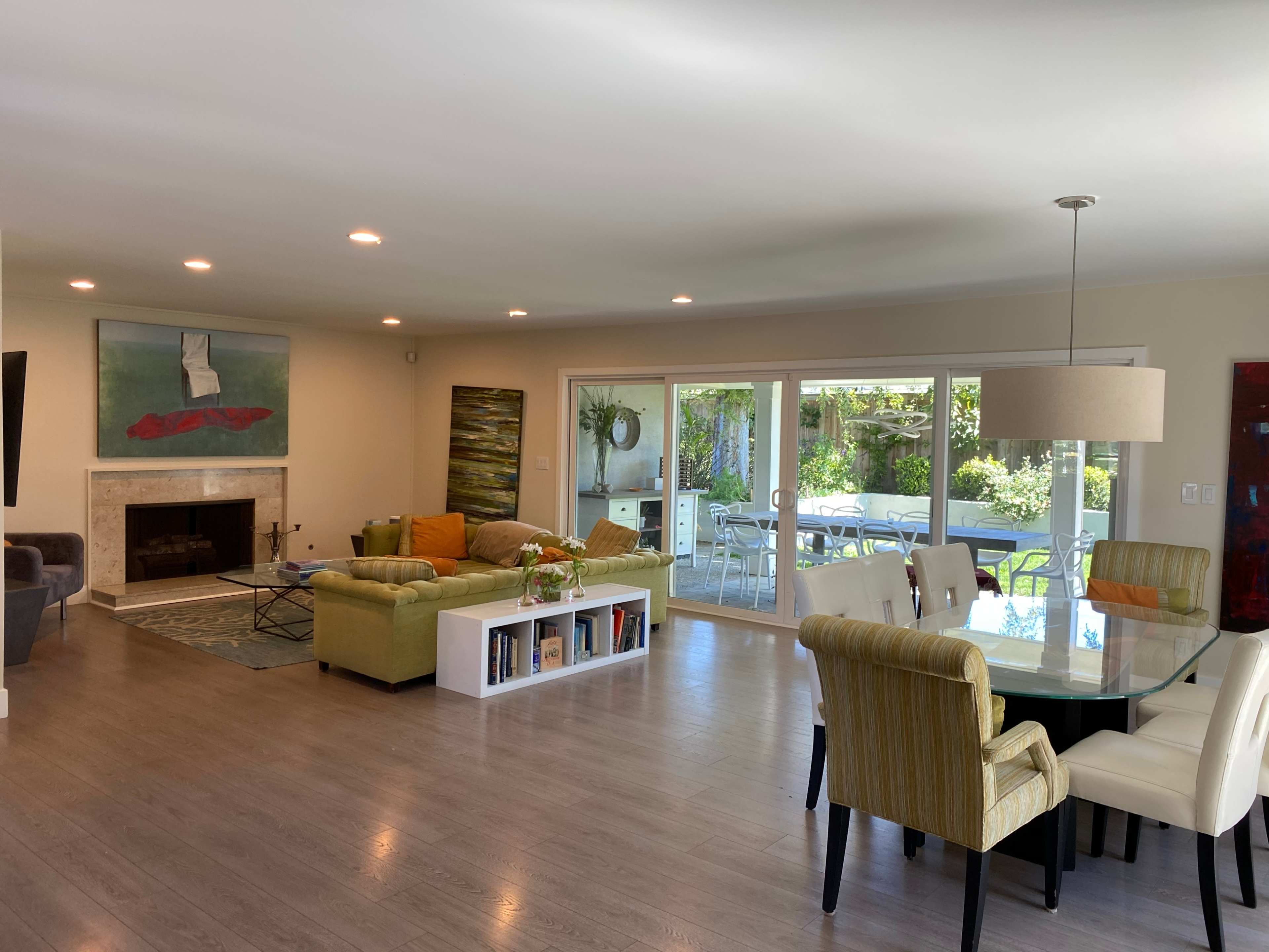 The image shows a spacious living area with a green sofa, a dining table, a fireplace, and large windows overlooking an outdoor patio.