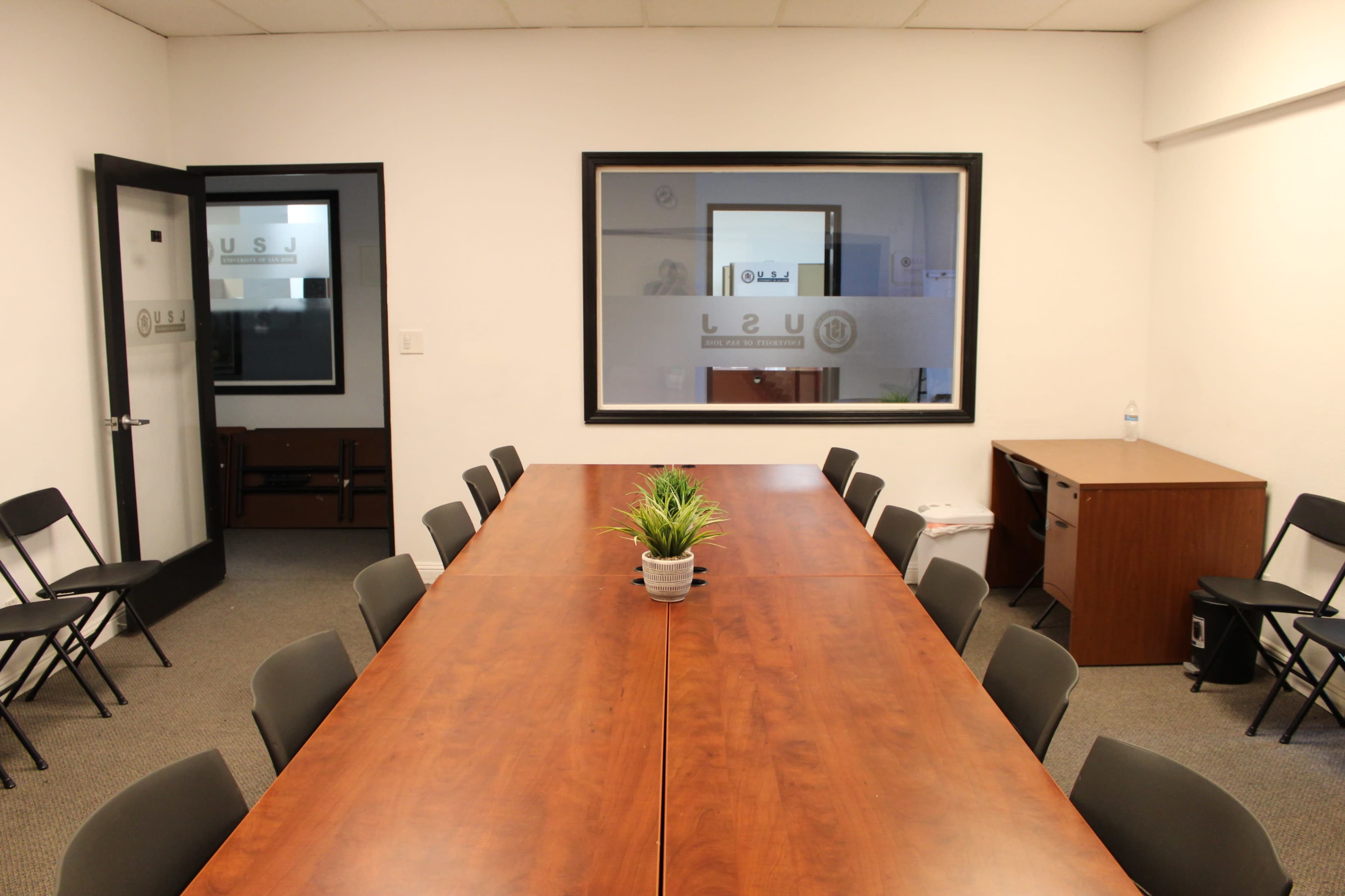 A long wooden table is centered in a conference room, surrounded by chairs, with a plant in the middle and a door leading to another room visible in the background.