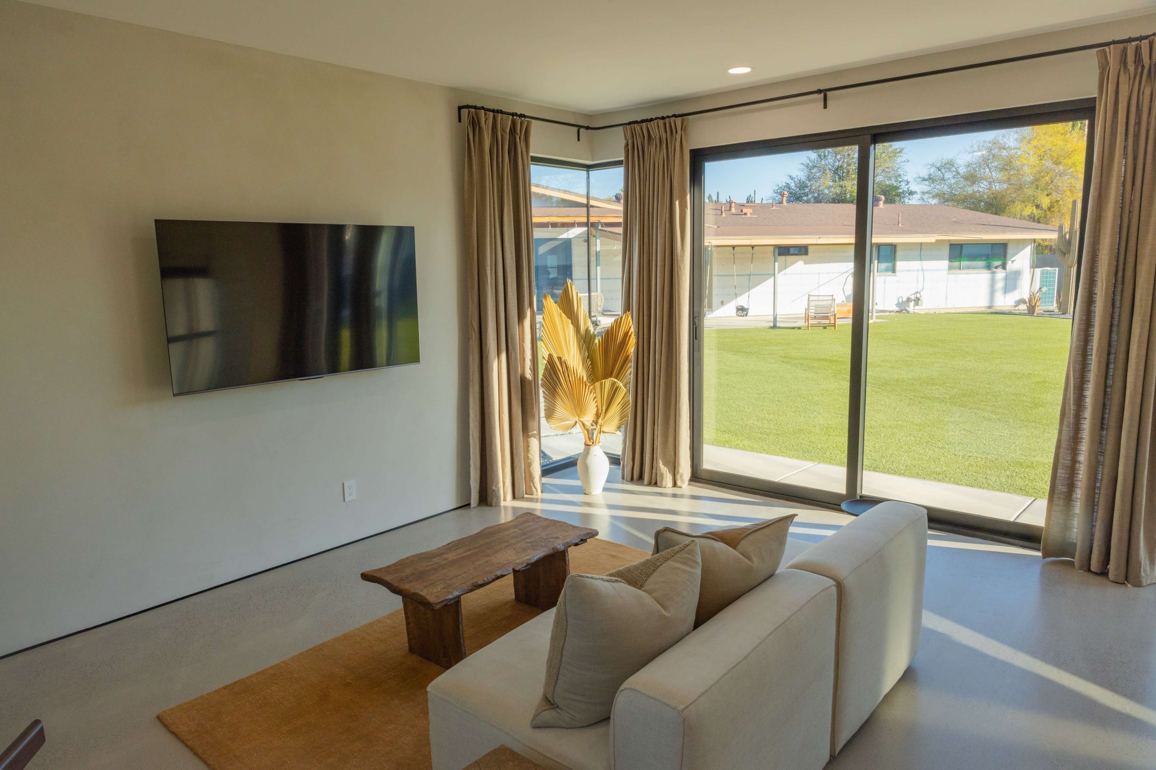 A modern living room features a TV mounted on a wall, a low wooden table, and a sofa with cushions, overlooking a backyard through large glass sliding doors.