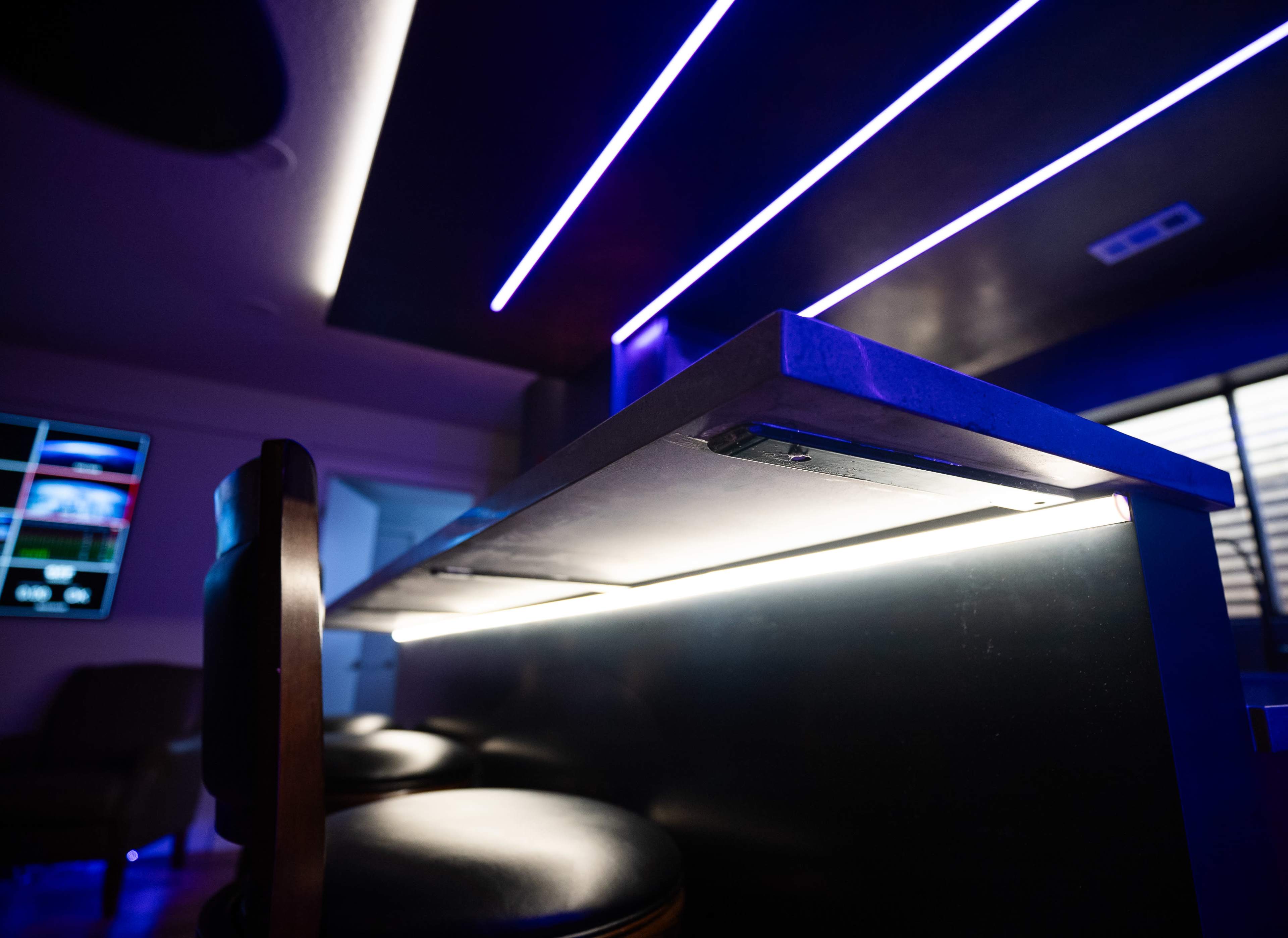 The image shows a modern bar counter with blue LED strip lighting, illuminated from below, and bar stools positioned in front.