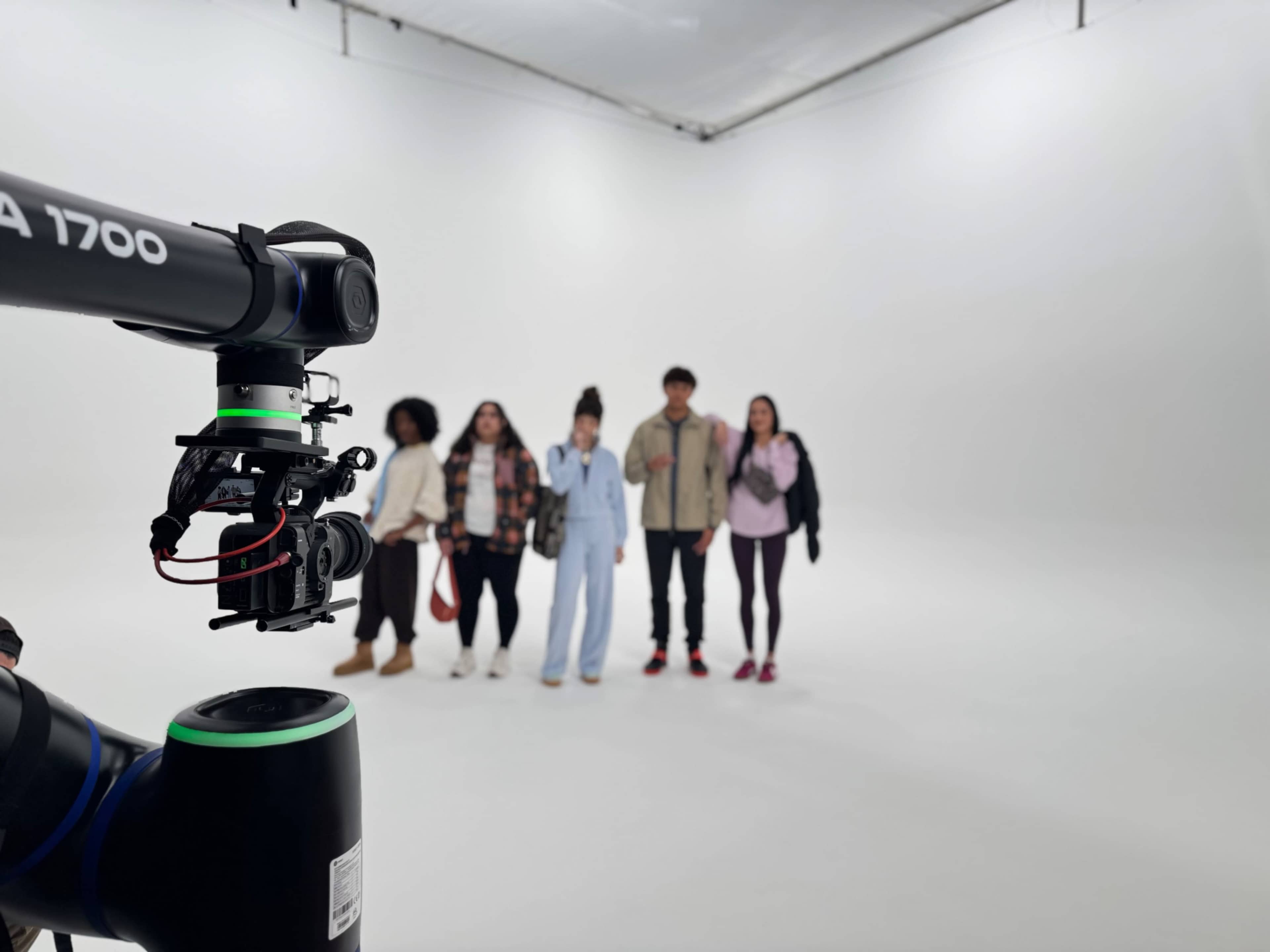 A camera on a tripod captures a group of five individuals standing together in a minimalist, well-lit studio space.