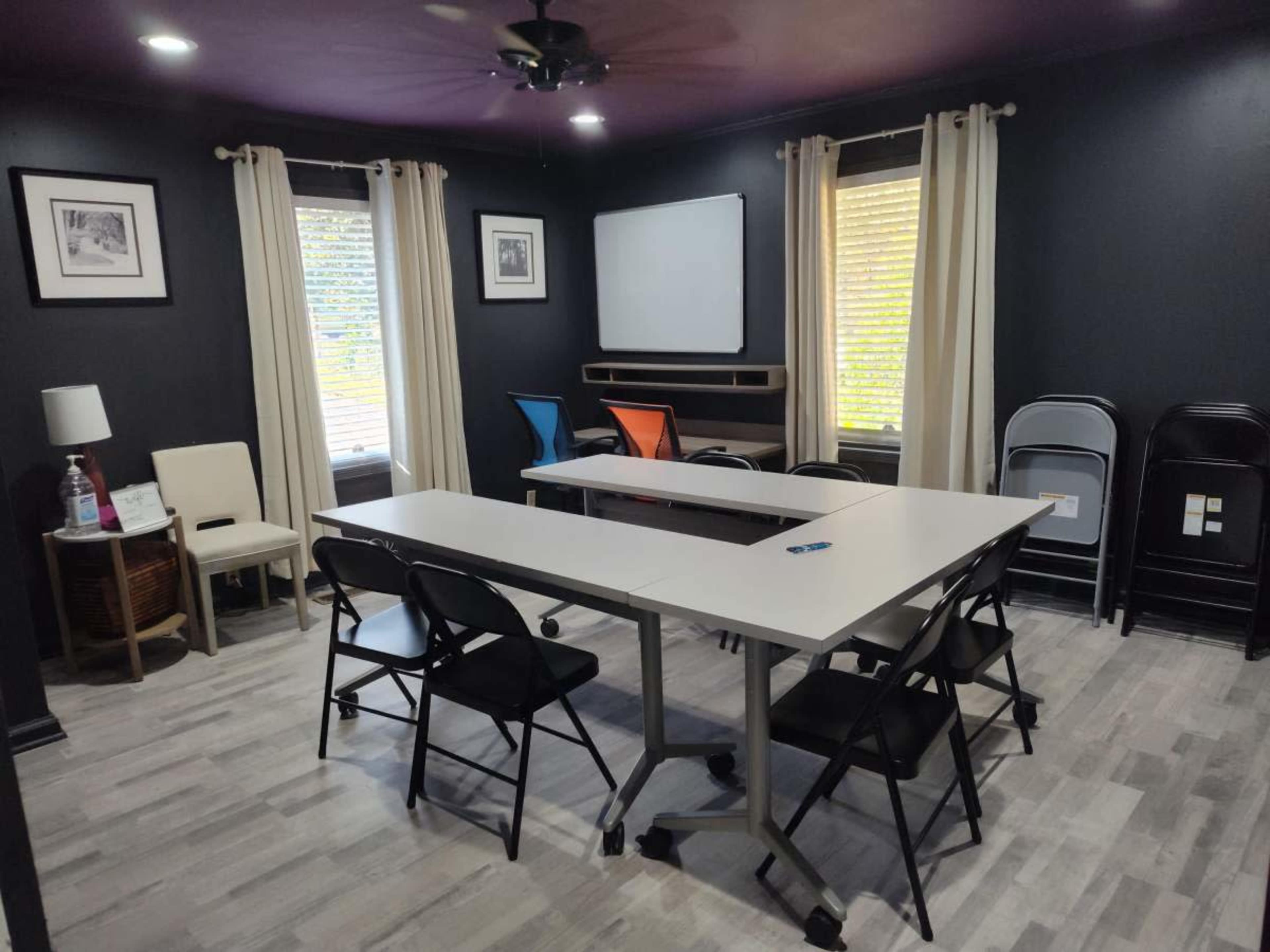 A small meeting room features a rectangular table surrounded by black folding chairs, with a projector screen mounted on the wall and windows letting in natural light.