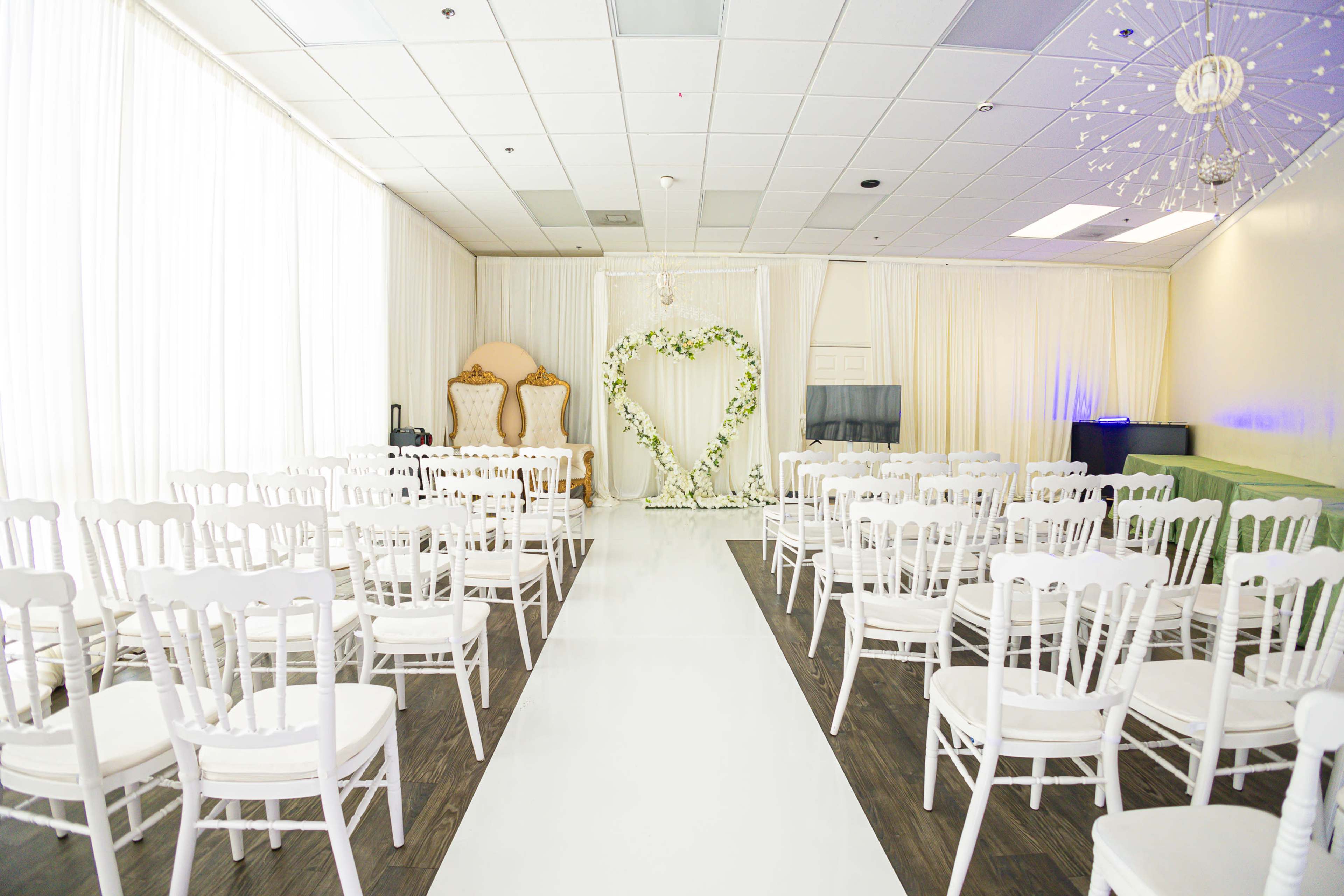 A wedding ceremony setup with rows of white chairs facing a floral heart arch in a bright room with sheer curtains.