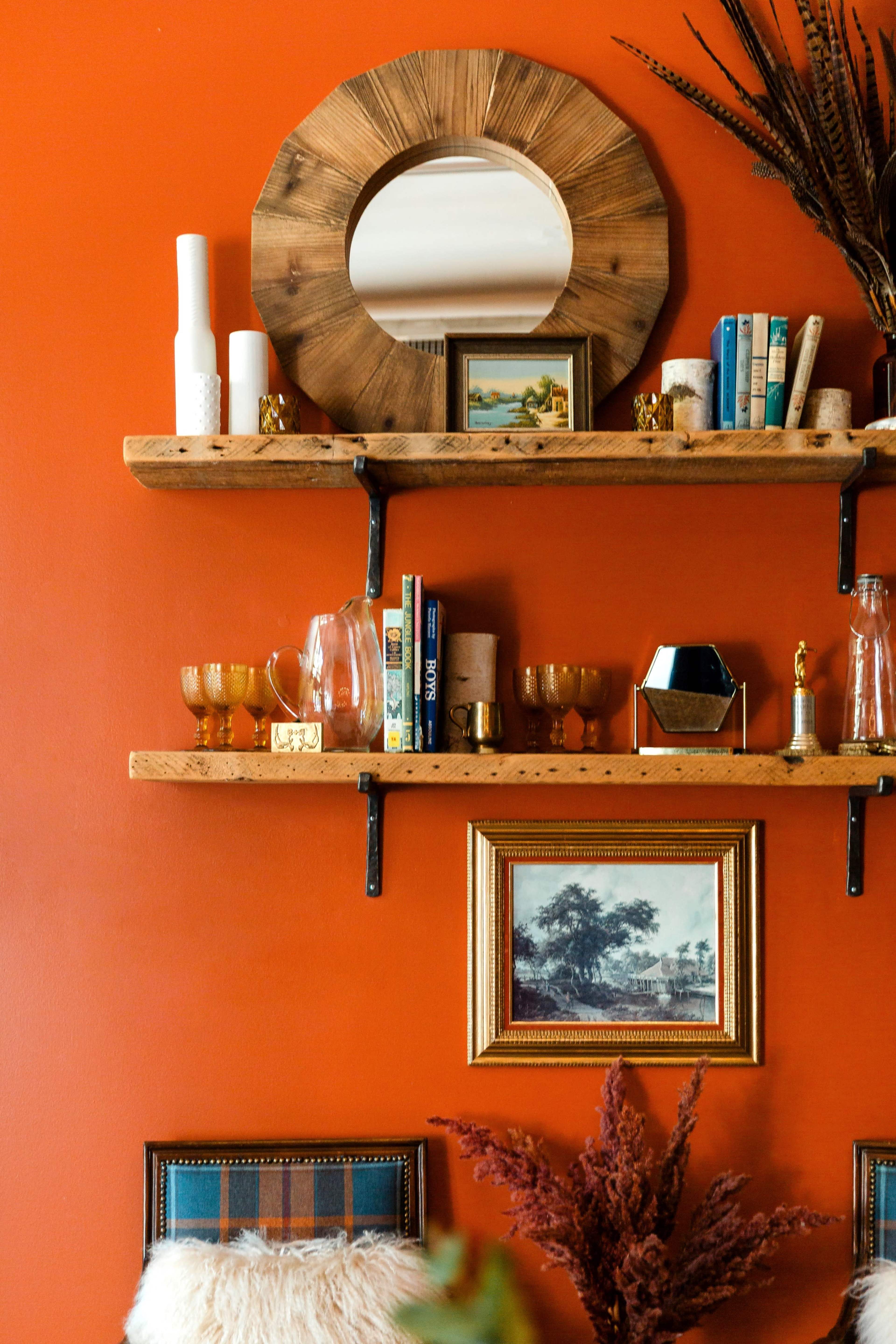 The image features a vibrant orange wall with two wooden shelves displaying various decorative items, including books, glasses, a framed painting, and a mirror.