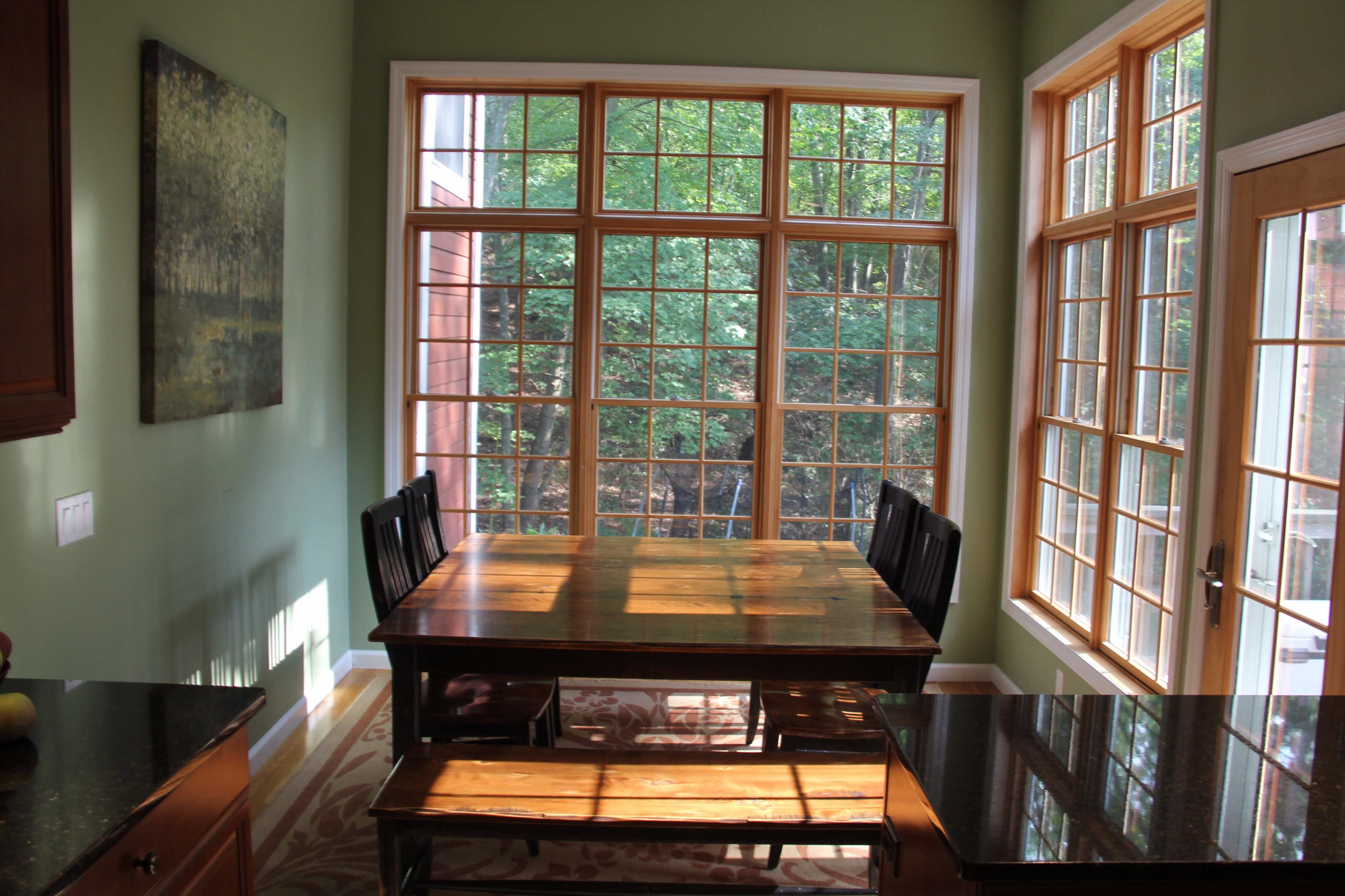 A wooden dining table with black chairs is positioned in front of large windows overlooking a green outdoor view.