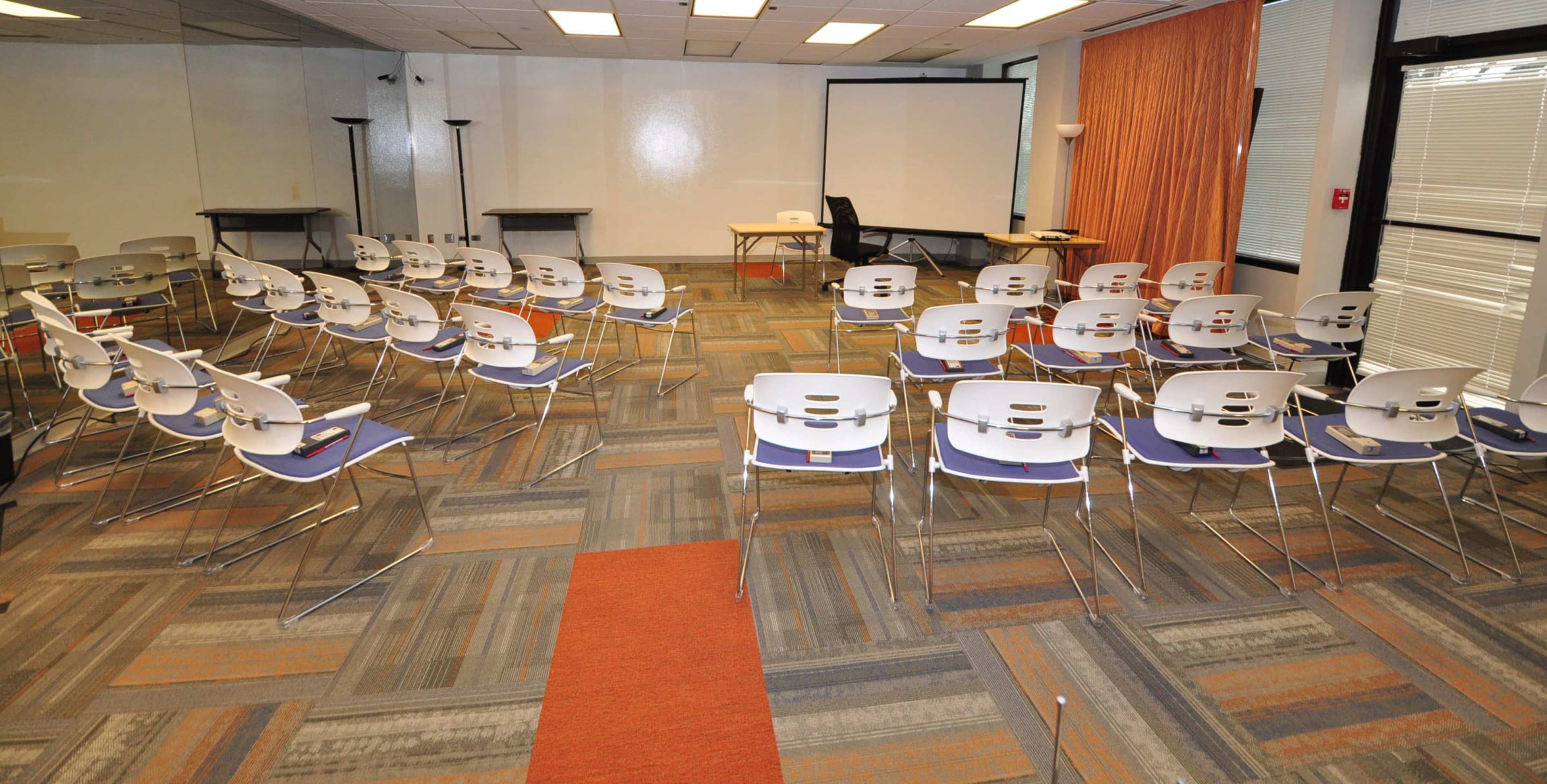 A room is set up with rows of chairs facing a blank screen and a table at the front.