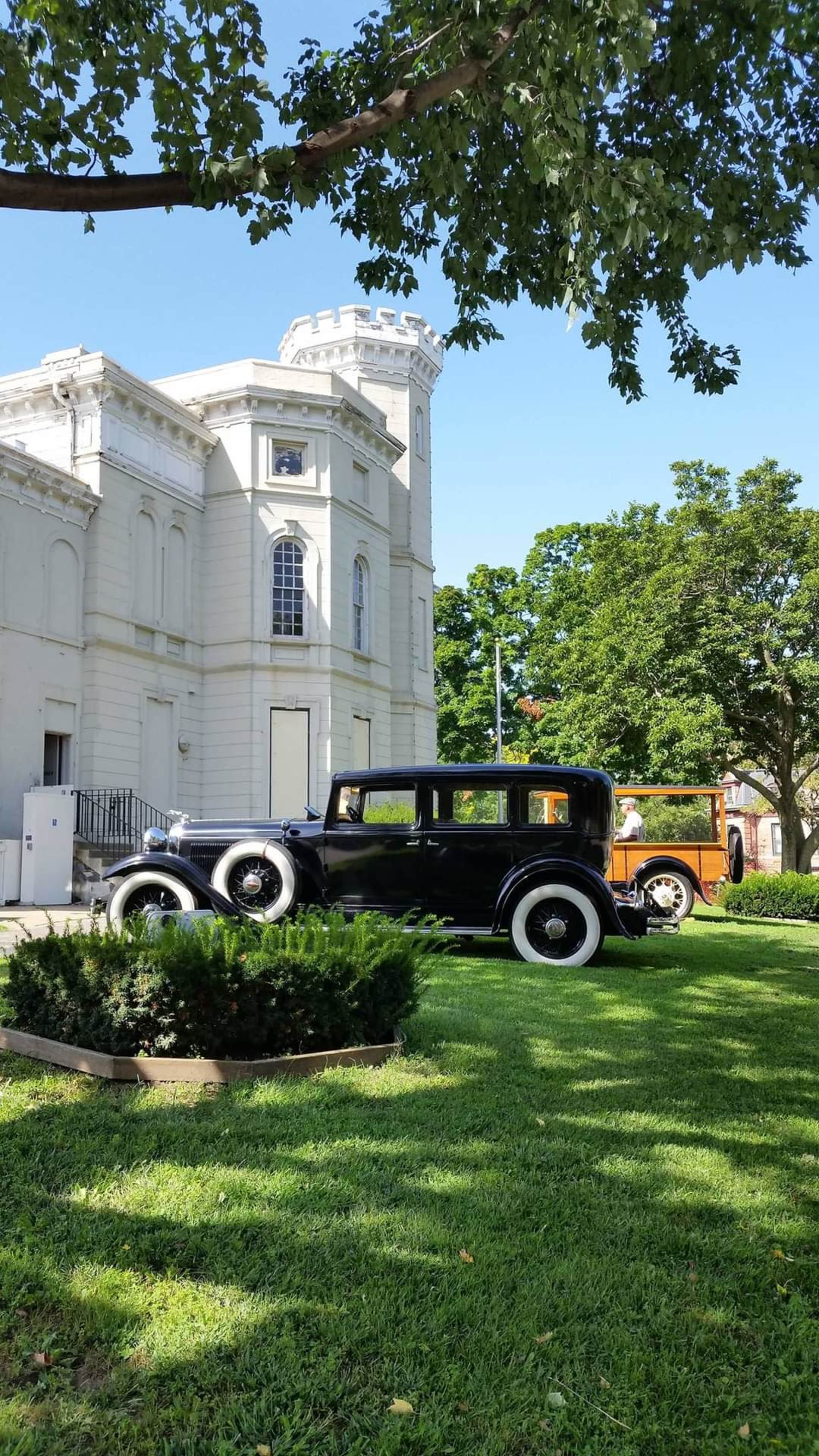 Two vintage cars are parked on a green lawn in front of an ornate white building under clear blue skies.