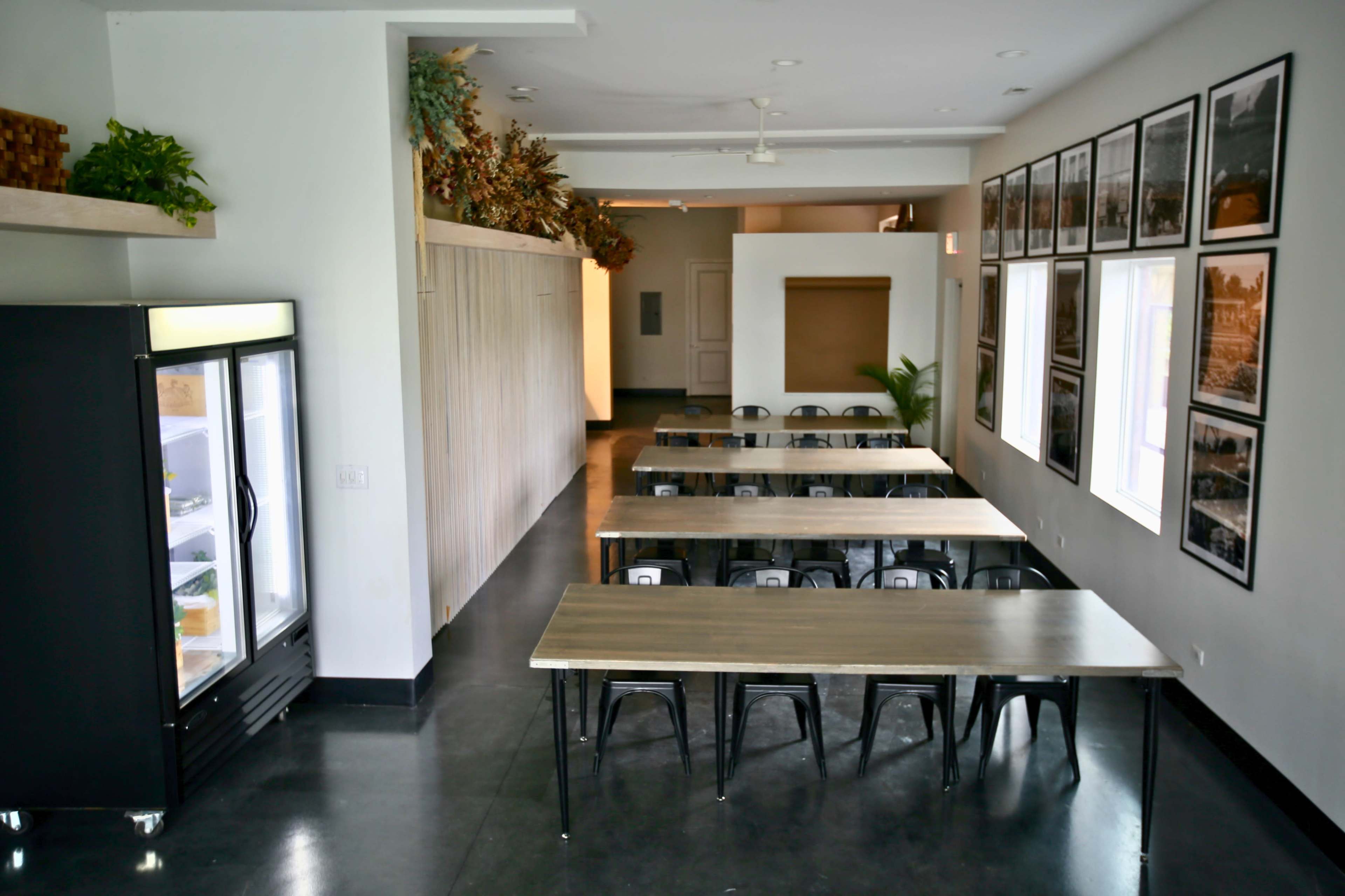 A spacious room with several long tables arranged in rows, a refrigerator in the corner, and framed photographs lining the walls.