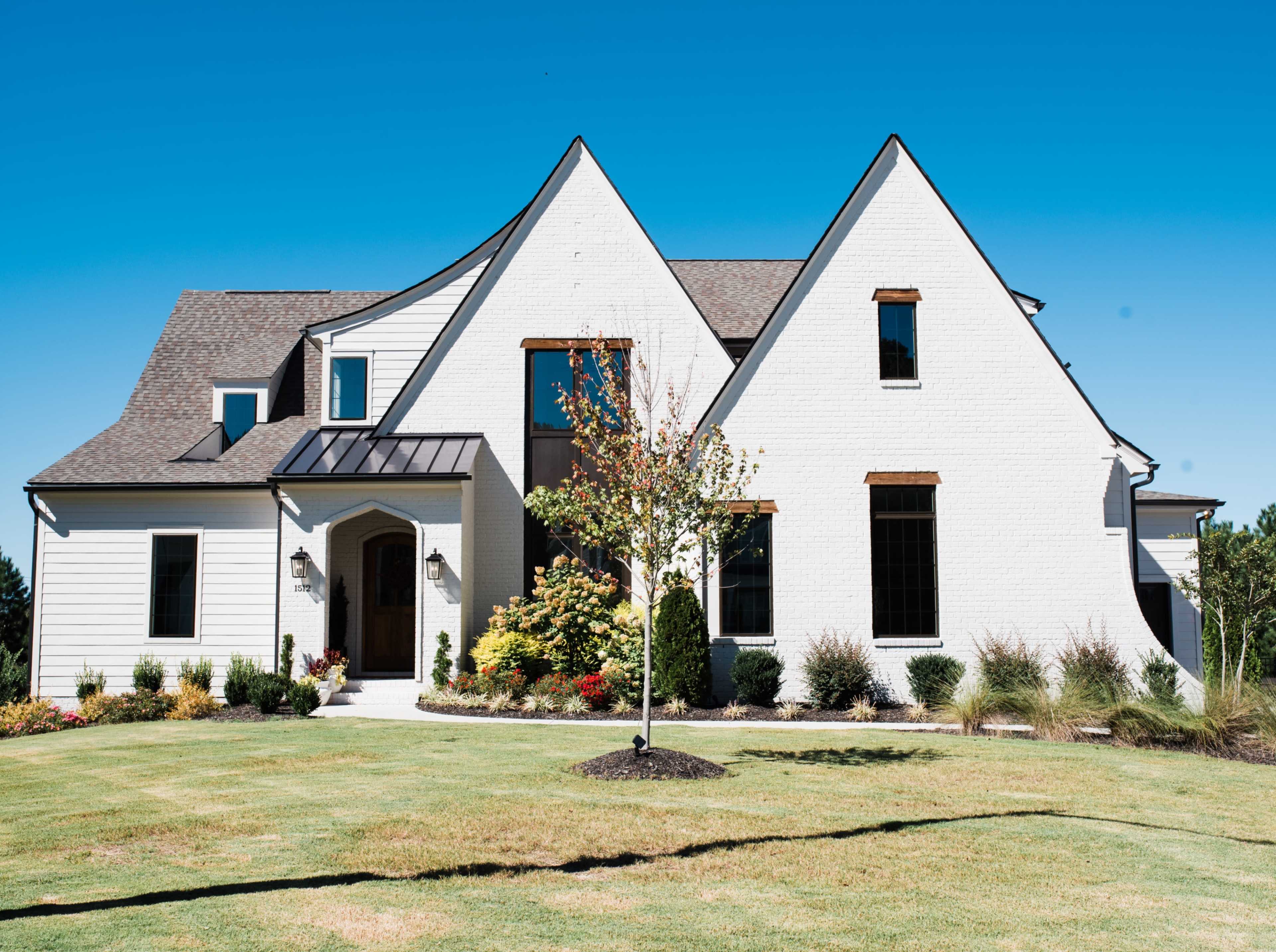 A modern white house with a distinctive roofline and landscaped front yard is shown under a clear blue sky.