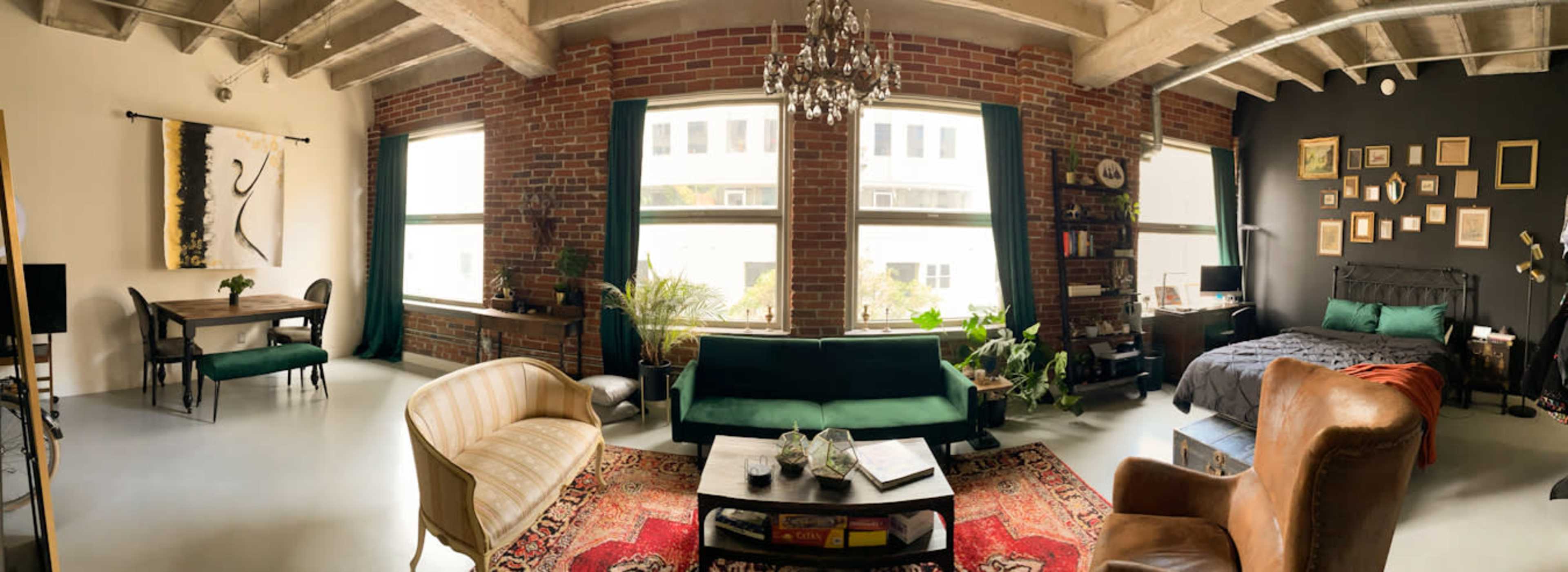 The image shows a spacious living area with large windows, exposed brick walls, various plants, and a mix of modern and vintage furniture.