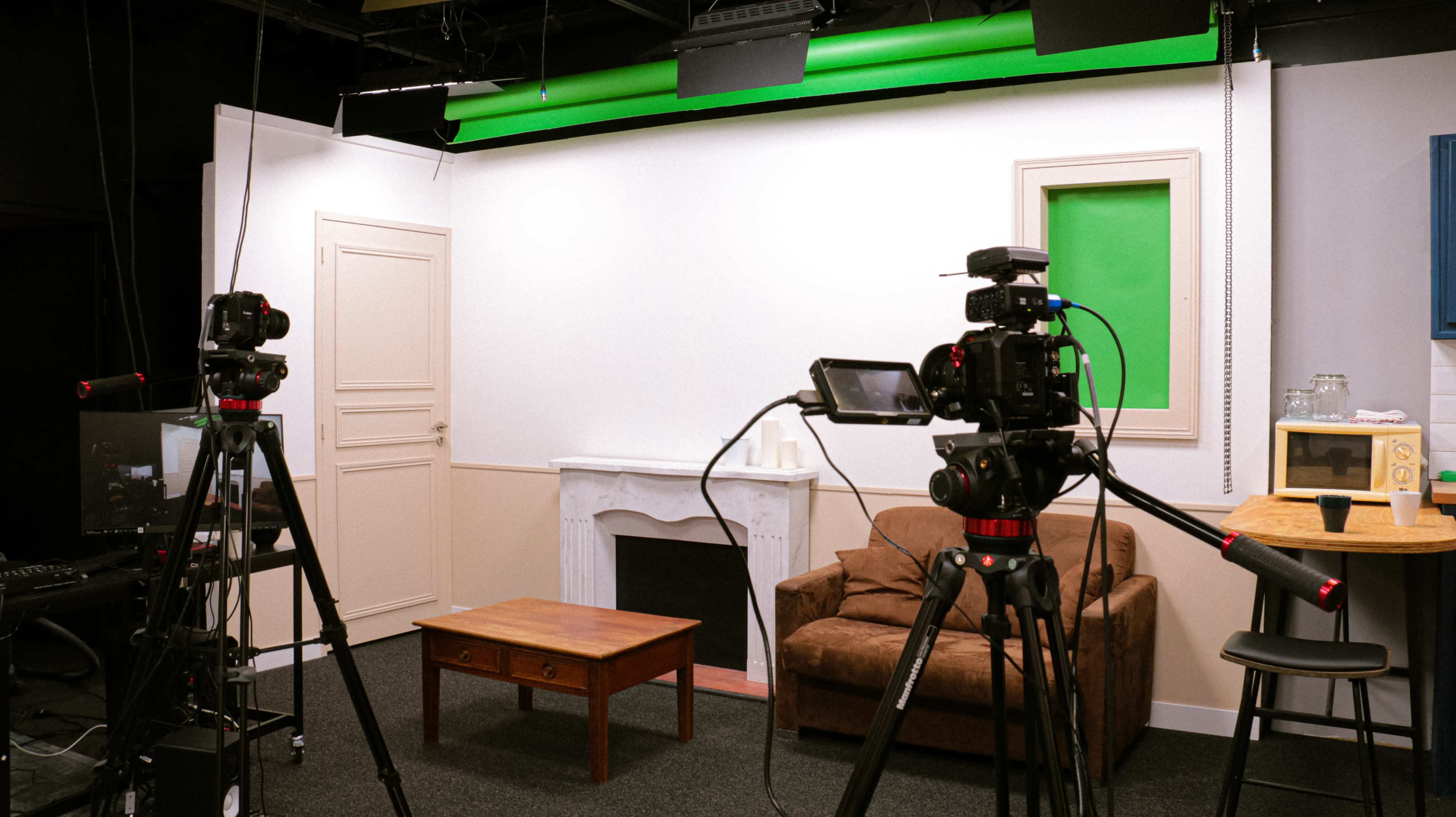 A studio setup with two cameras focused on a small living space featuring a brown couch, a wooden coffee table, and a green screen on the wall.